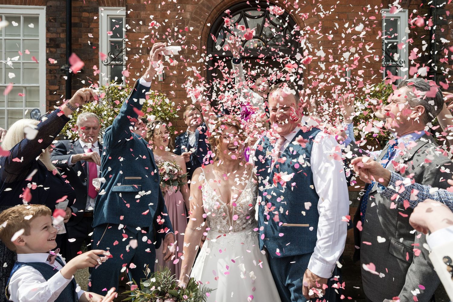 Bride and groom walking down Kelham House steps as guests throw confetti, with the manor house behind.