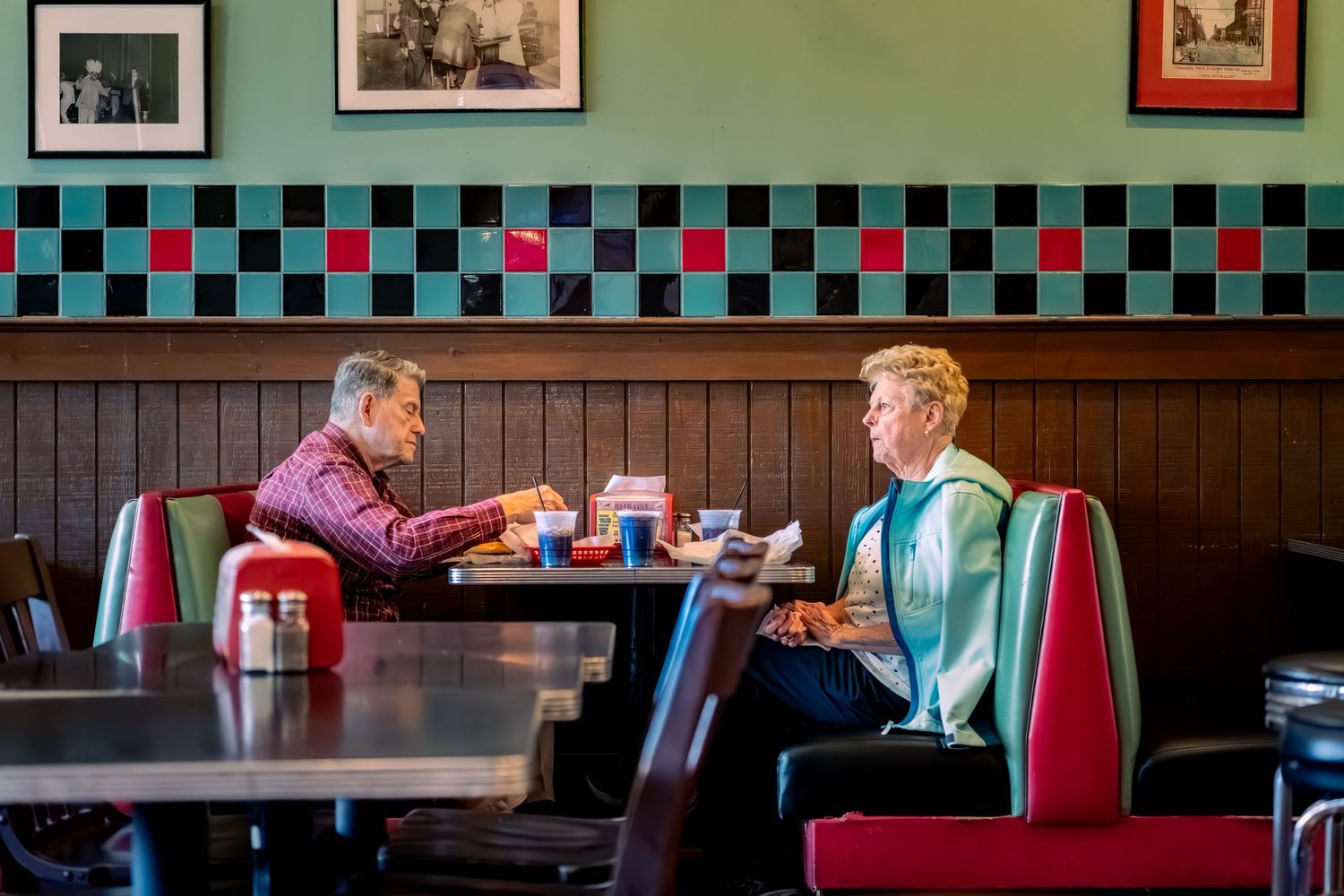 A retro diner interior with red vinyl booths, checkered walls, and vintage photographs displayed in frames.