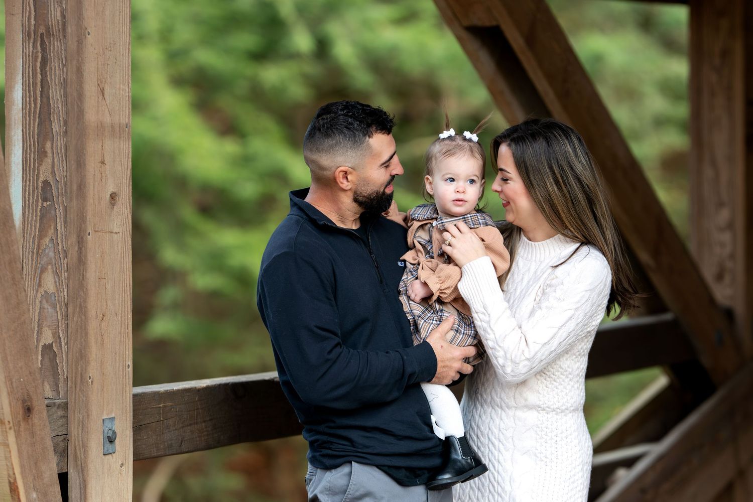 Young couple holds their baby girl on a wooden covered bridge surrounded by lush green trees.