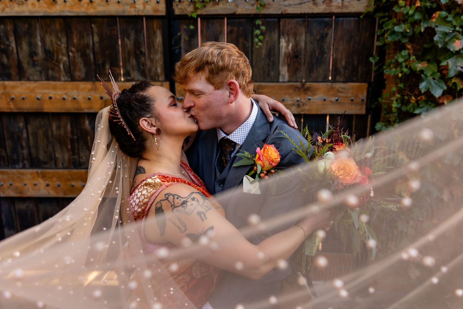 Bride and groom sharing a romantic kiss at their wedding, with a flowing veil in the foreground and rustic wooden backdrop.