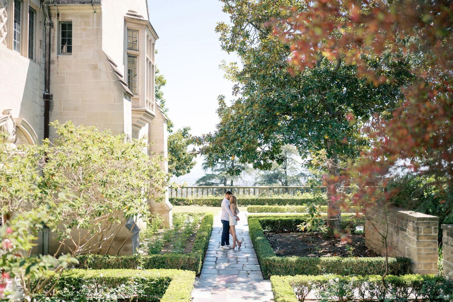Couple strolling through elegant formal garden pathway beside historic mansion surrounded by lush greenery.