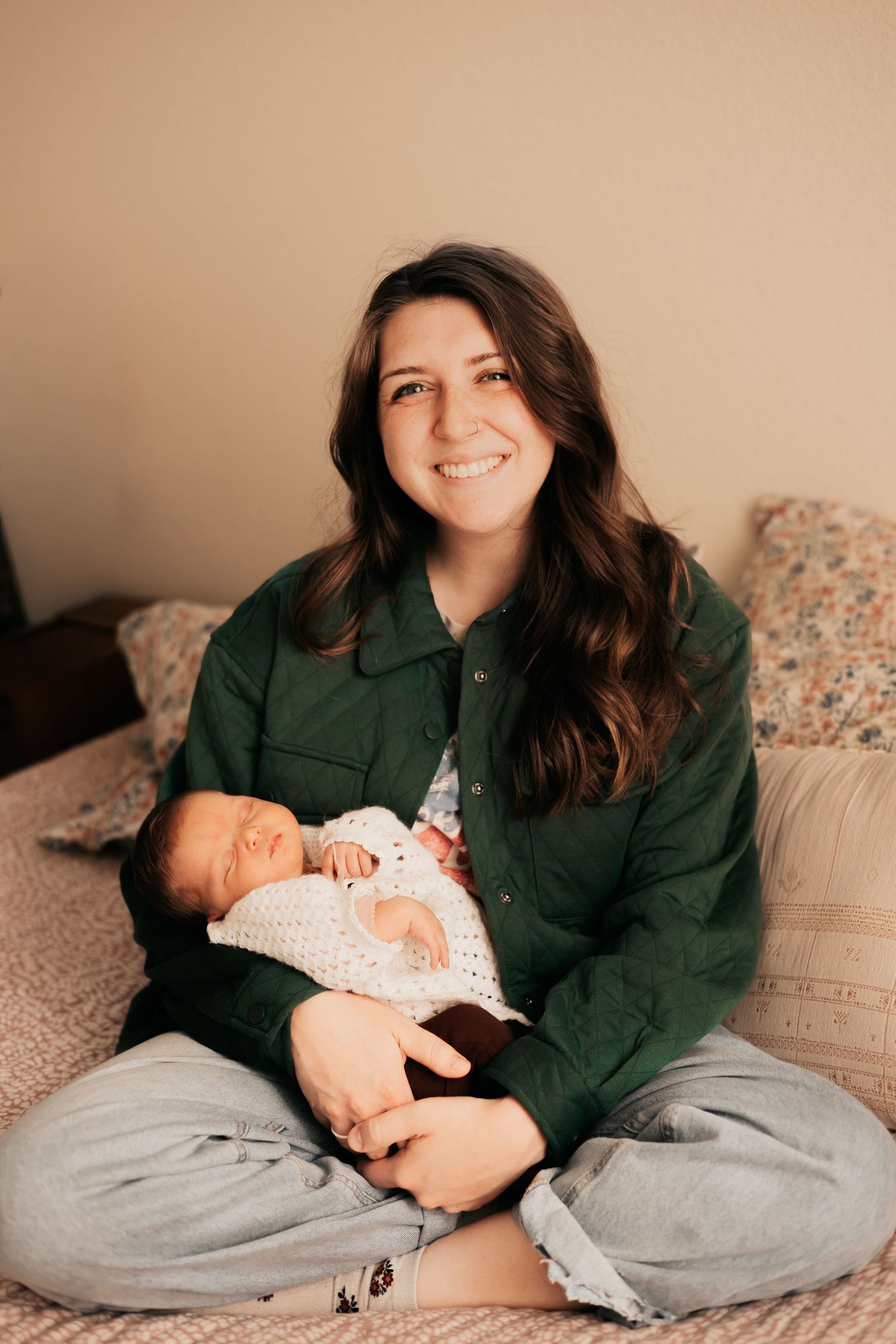 Smiling young woman in green jacket sitting cross-legged on bed holding sleeping newborn | Newborn Photos When You Don't Feel Like Yourself