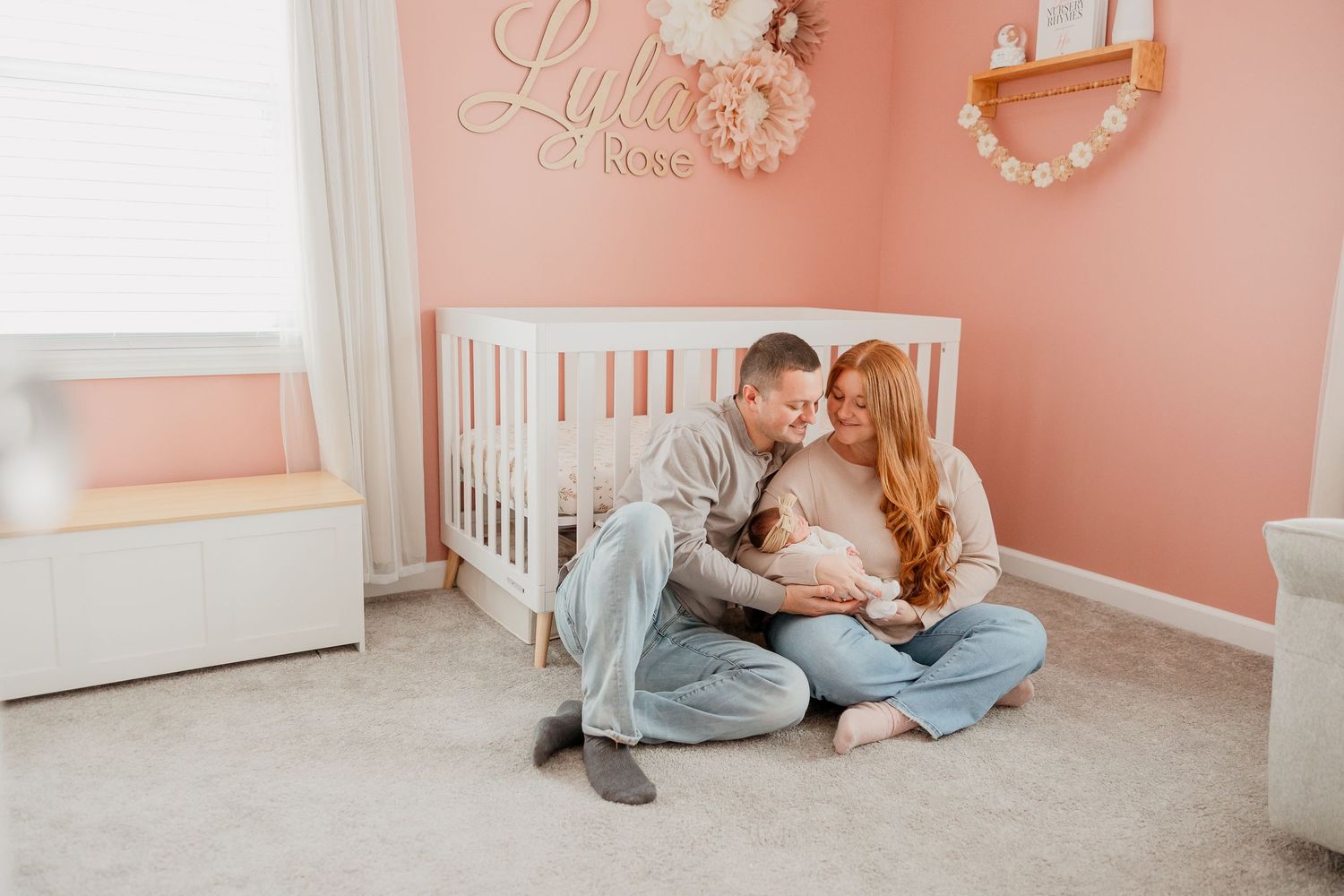 Mom and dad sit on the nursery floor holding their newborn daughter beside her crib.