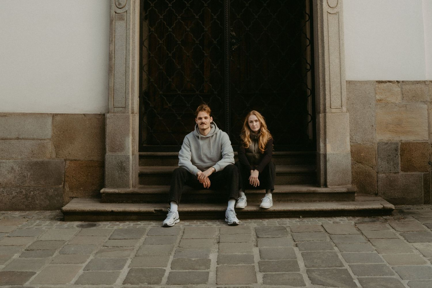 Two people sit together on stone steps in front of a large wooden door wearing casual clothing.