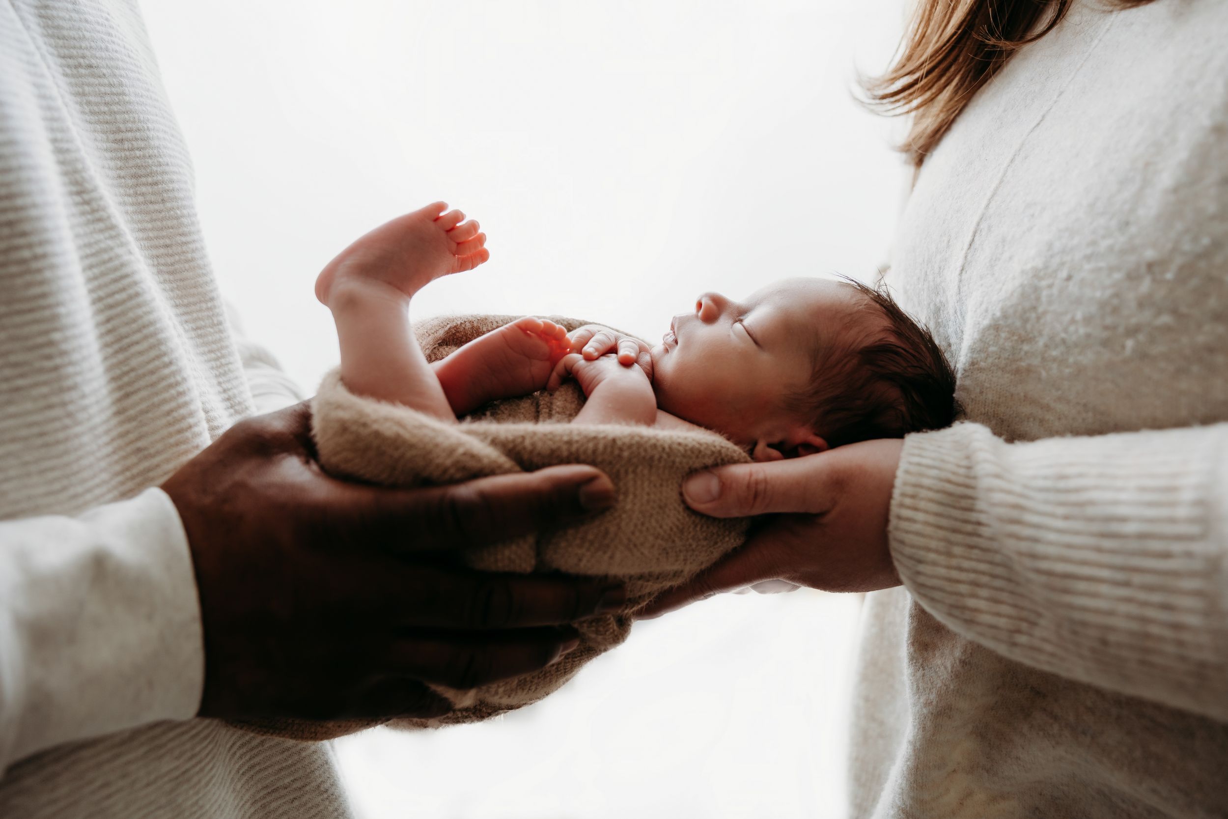 A newborn baby lies between two people wearing white sweaters holding the infant gently.