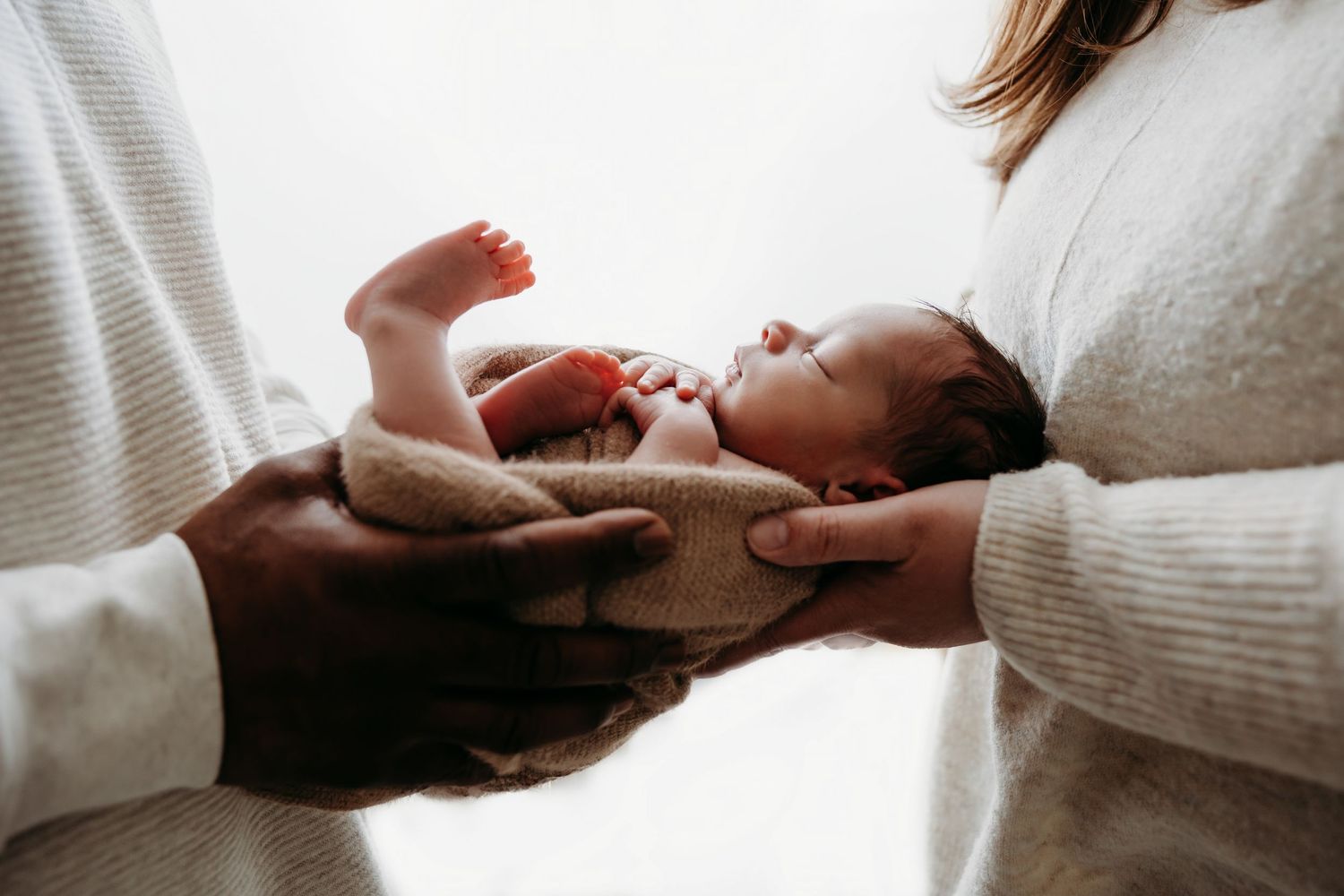 A newborn baby lies between two people wearing white sweaters holding the infant gently.