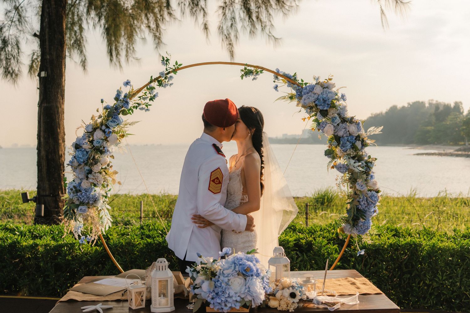 A couple shares a romantic moment under a floral arch by a serene lakeside during golden hour.