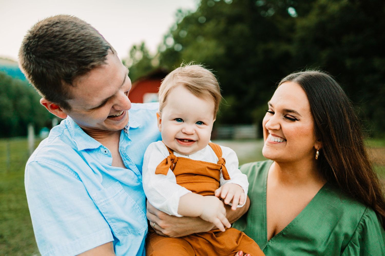 Happy young family with smiling baby in rust overalls outdoors near red barn on sunny day.