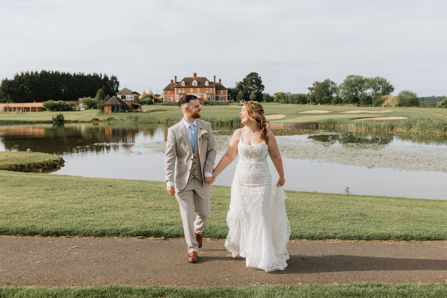 Bride and groom walk hand-in-hand on a path beside a serene pond with a grand estate in the background.