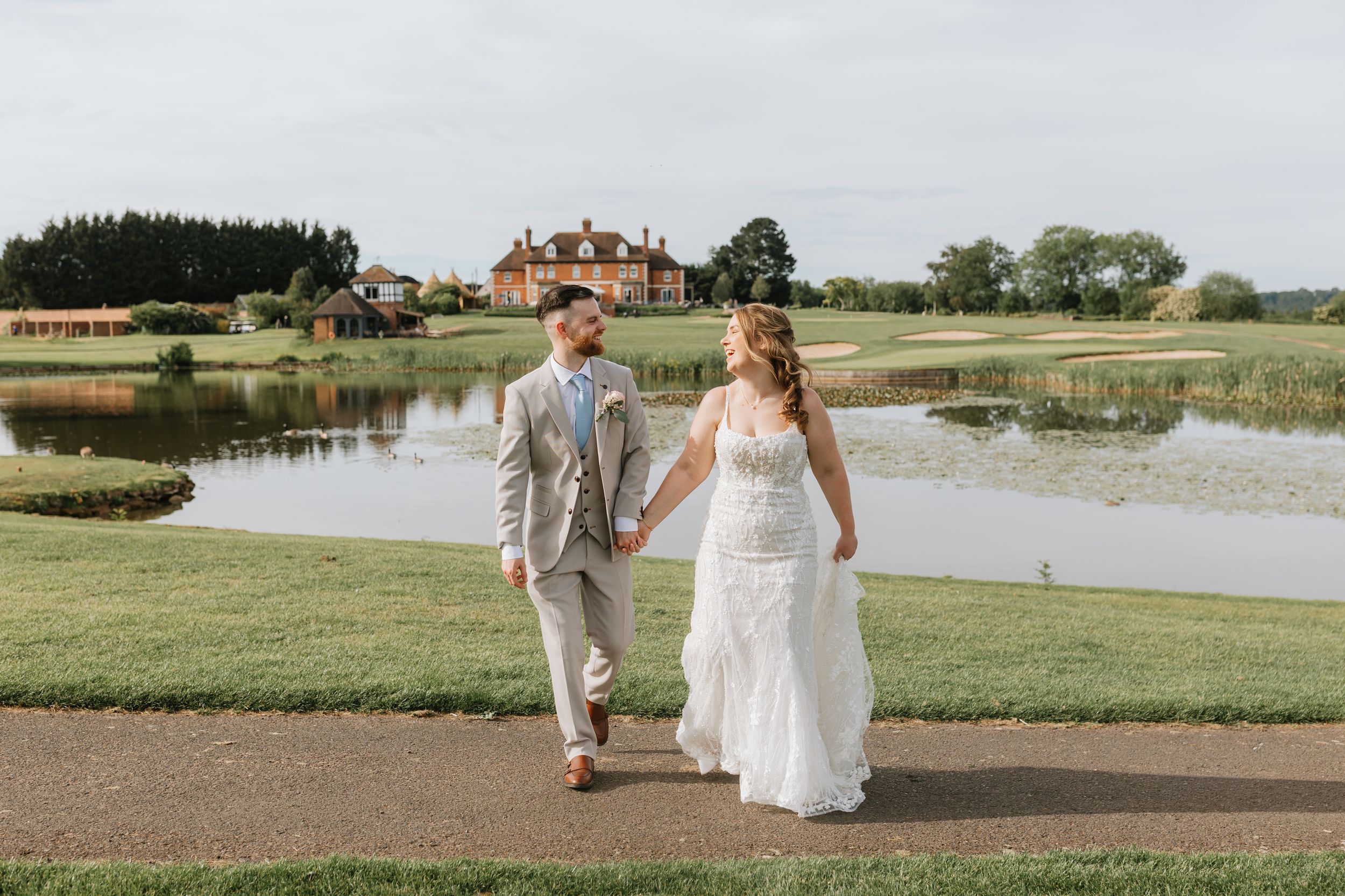 Bride and groom walk hand-in-hand on a path beside a serene pond with a grand estate in the background.
