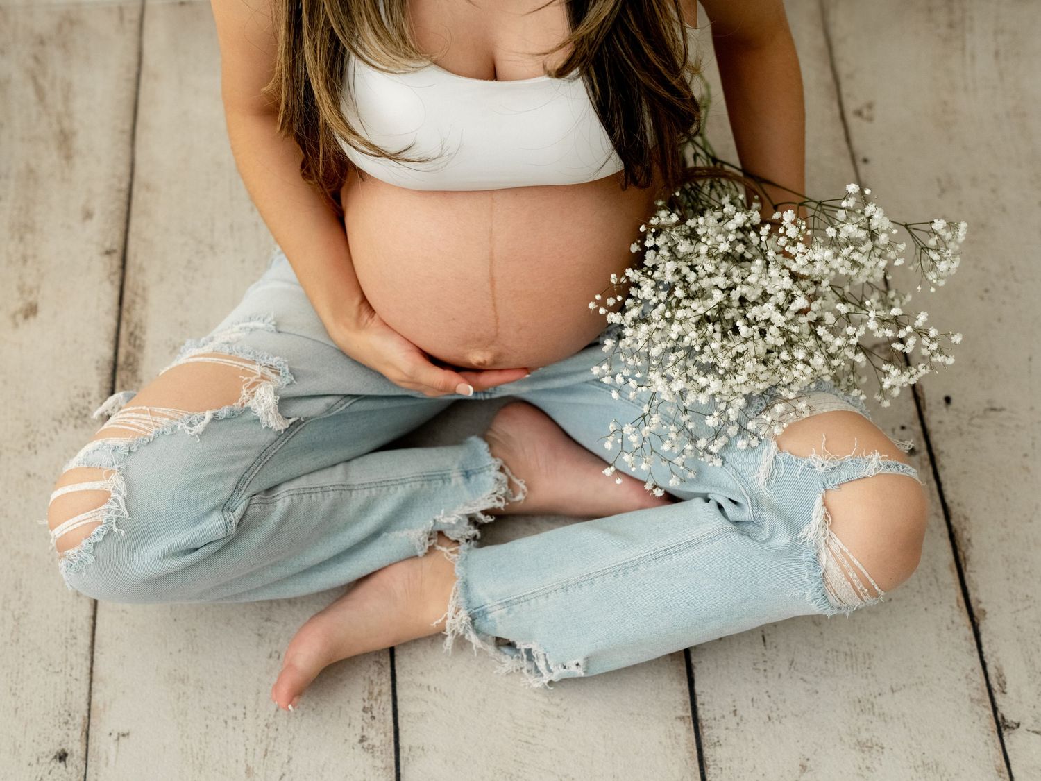 Pregnant woman in white crop top and ripped jeans cradling baby bump against white wall in studio maternity photo.