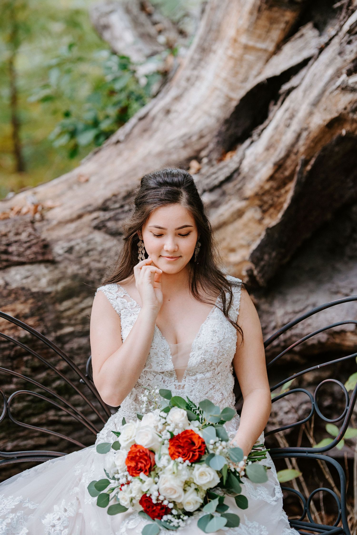 A bride in a white lace dress holds a red and white bouquet while sitting near a rustic wooden backdrop in nature.