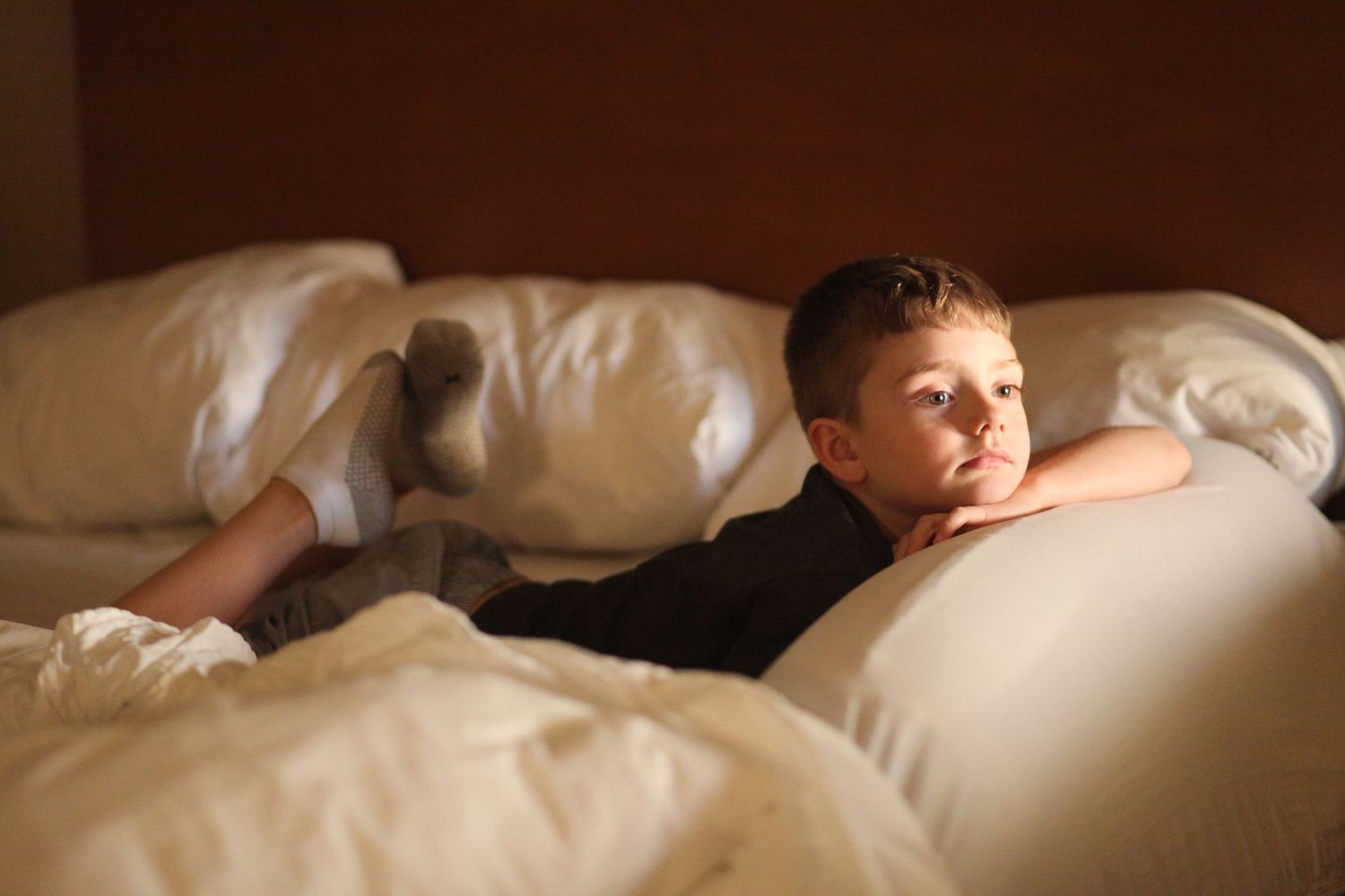 Young boy relaxing on white hotel bed pillows in dimly lit room.