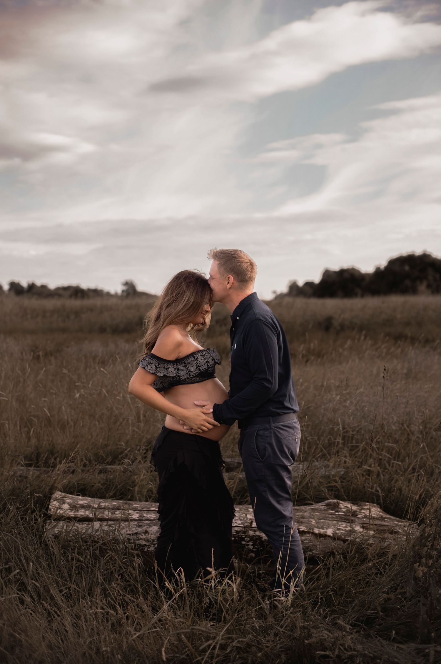 ett par står på en äng. kvinnan är gravid, de står mot varandra och mannen håller om magen och pussar kvinnan på pannan.