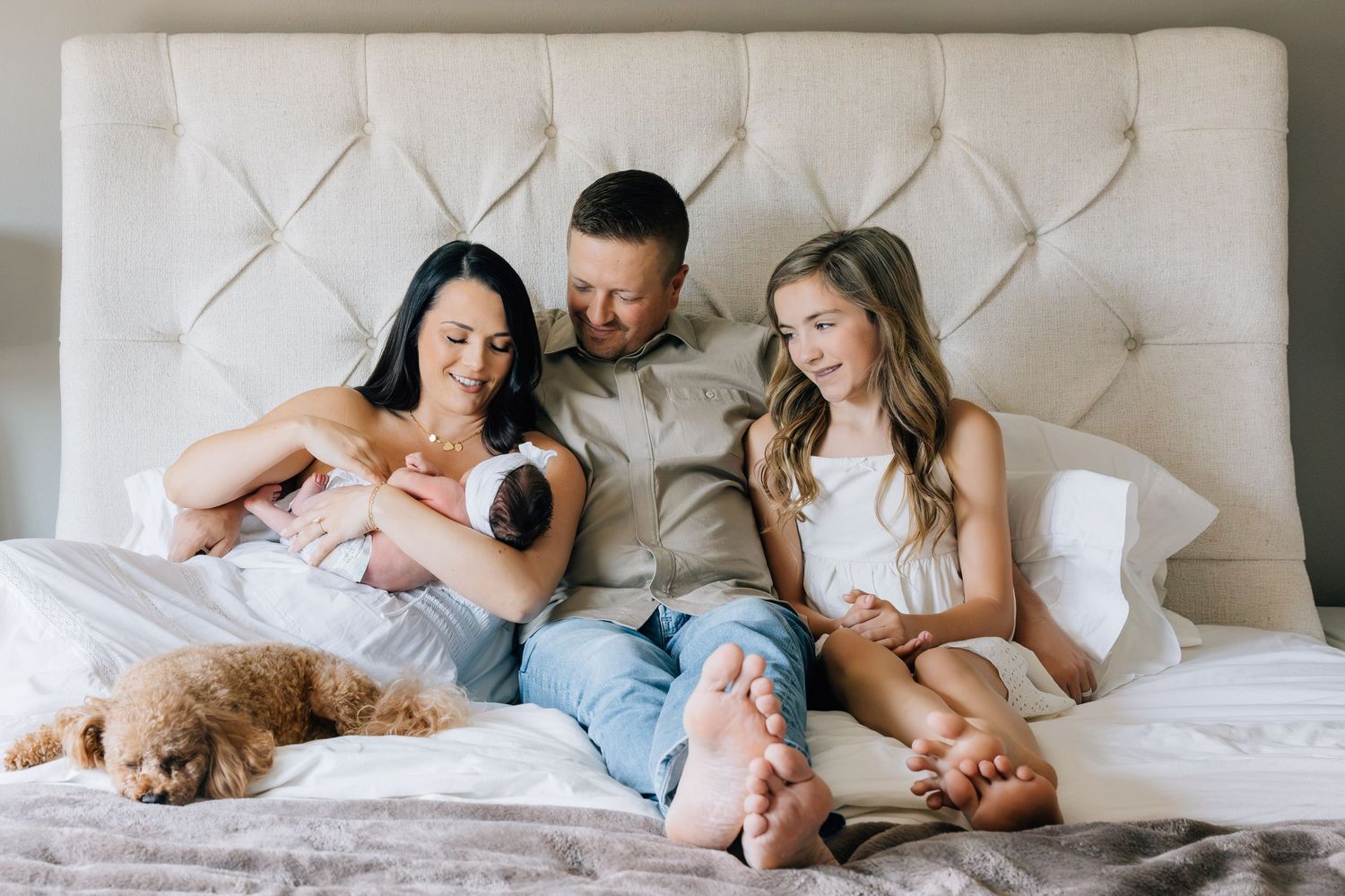 Family with newborn baby and brown poodle puppy relaxing together on a white bed at home.