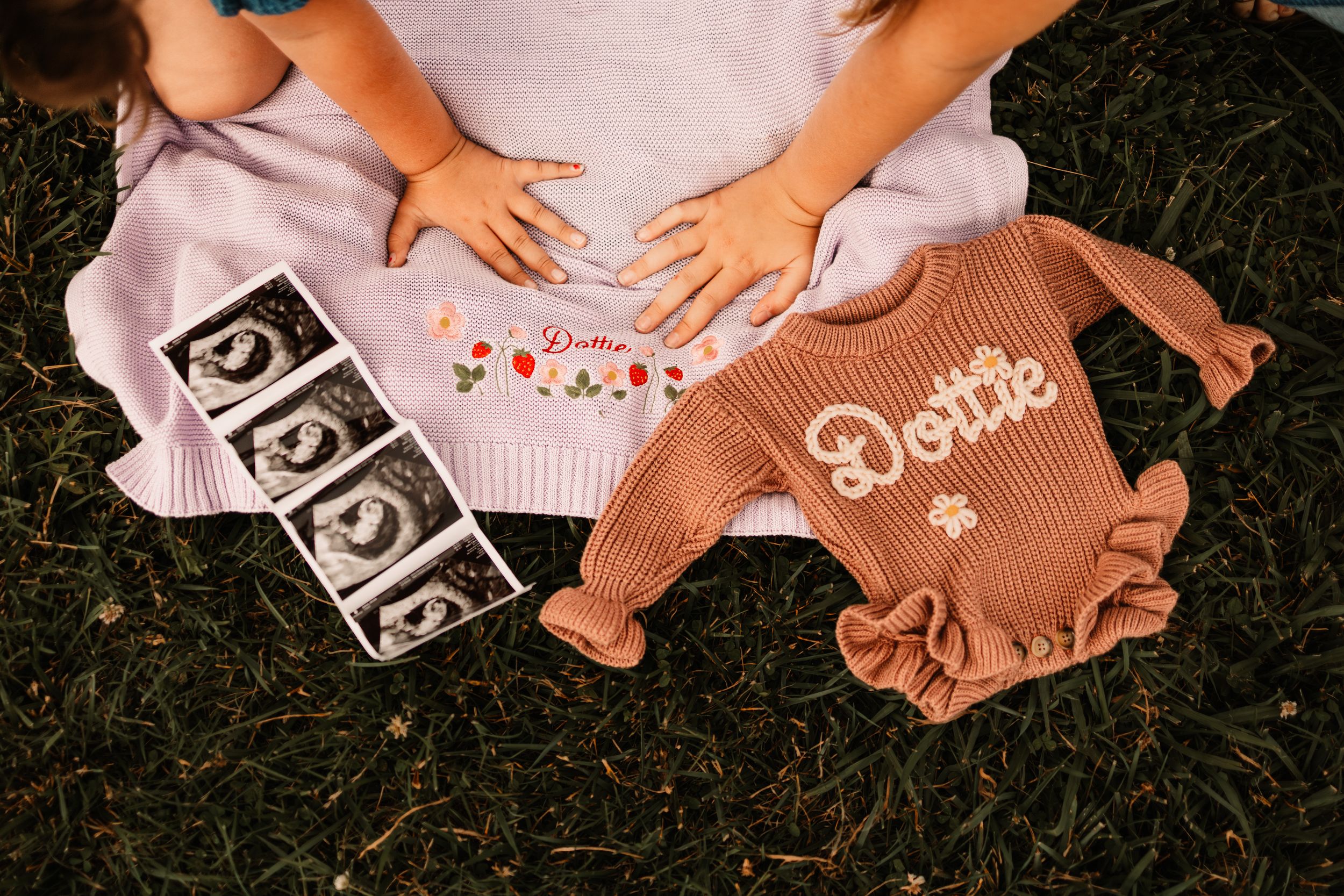 Pregnant woman holding ultrasound photos beside a brown ruffle romper with Dottie embroidered on it on green grass.