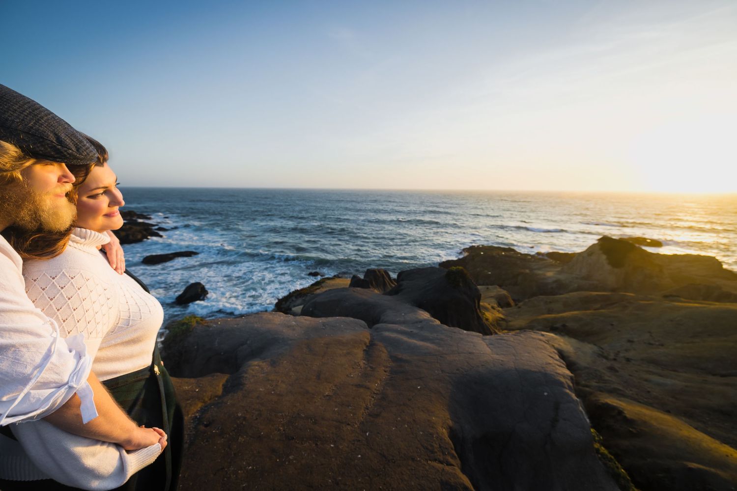 Couple embracing on rocky coastal cliffs watching a golden sunset over the ocean waves.