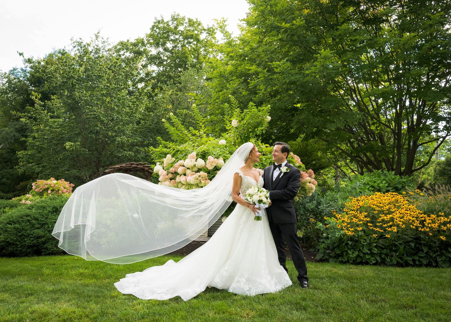 Bride and groom pose in lush garden, her veil flowing dramatically in the wind, surrounded by colorful blooms.