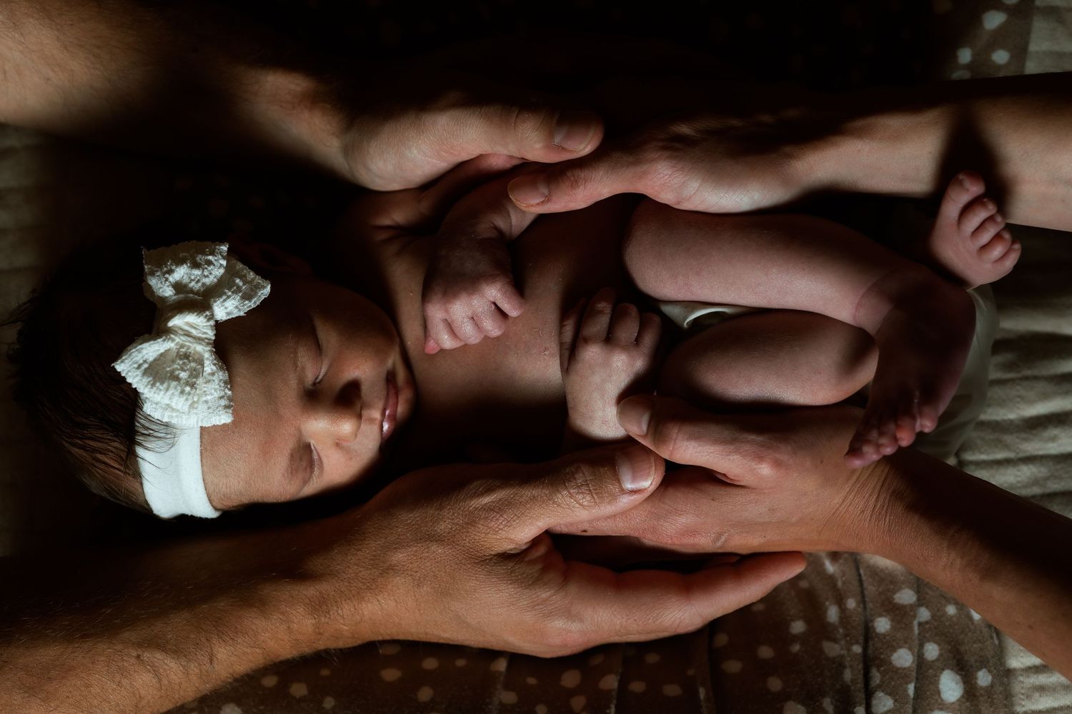 Newborn baby girl with white bow headband cradled in parents hands in soft light photography.