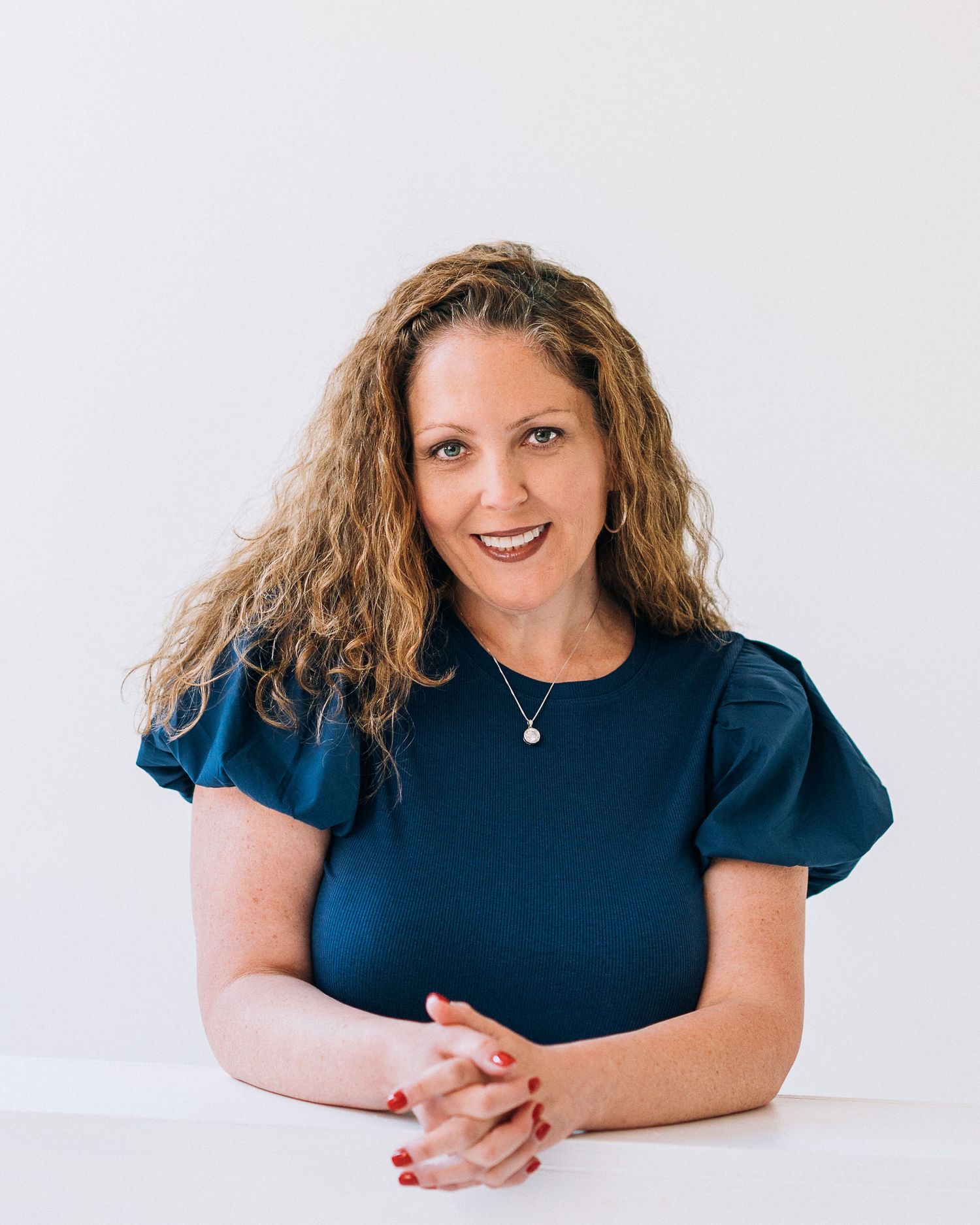 Professional woman with curly hair in a teal top, smiling confidently while seated at a white table.