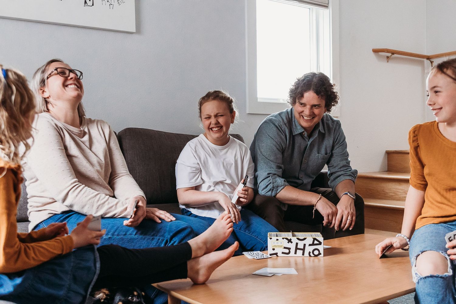 Group of five people laughing and playing a board game together in a cozy living room setting.