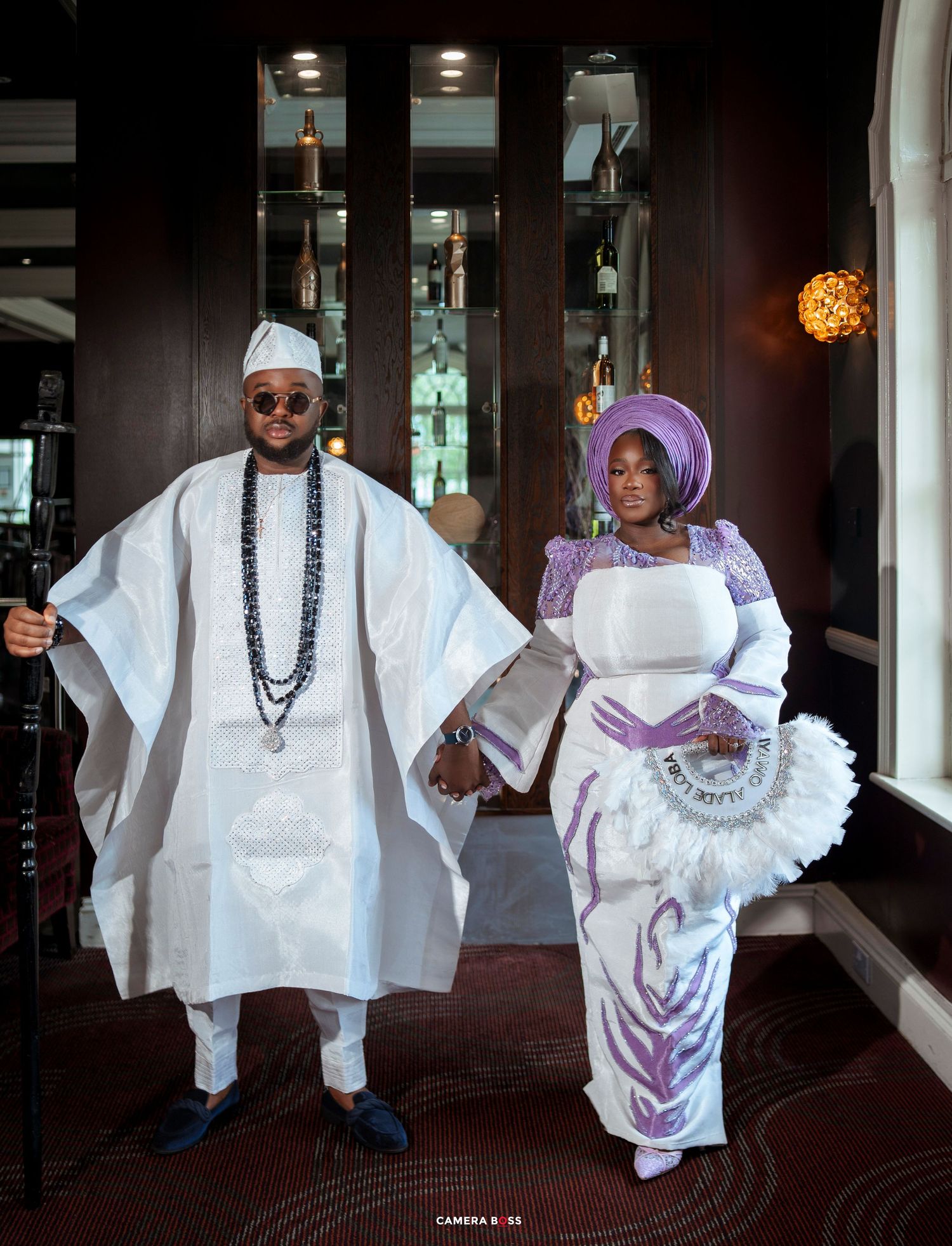 Nigerian couple in traditional Yoruba attire, man in white agbada and woman in purple and white iro and gele.