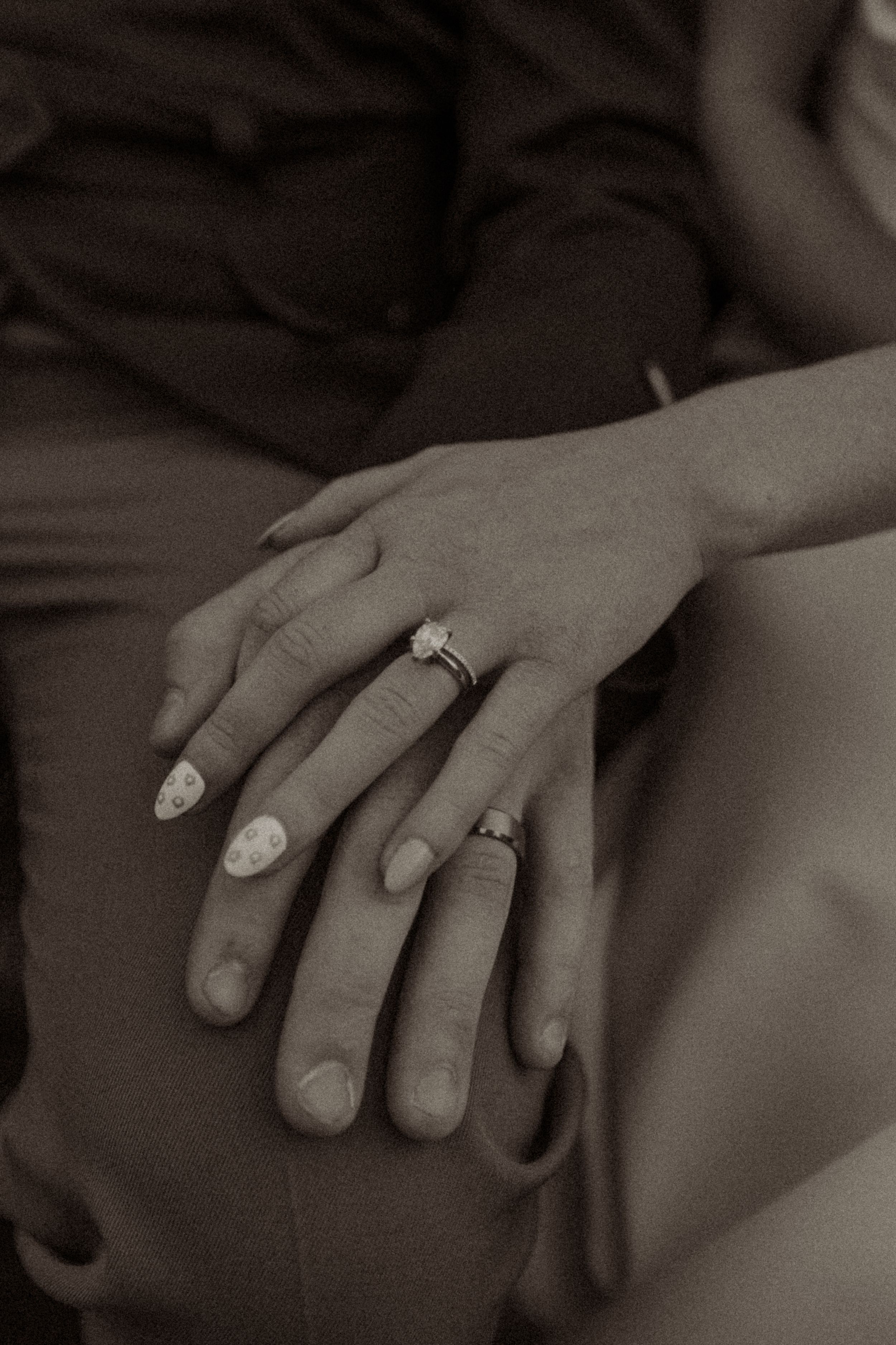 Sepia-toned close-up of two hands resting together, one wearing a diamond engagement ring and decorated nails.
