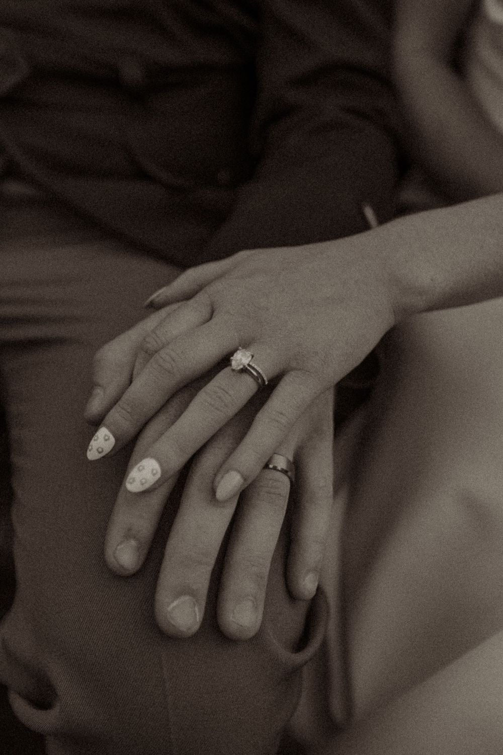 Sepia-toned close-up of two hands resting together, one wearing a diamond engagement ring and decorated nails.