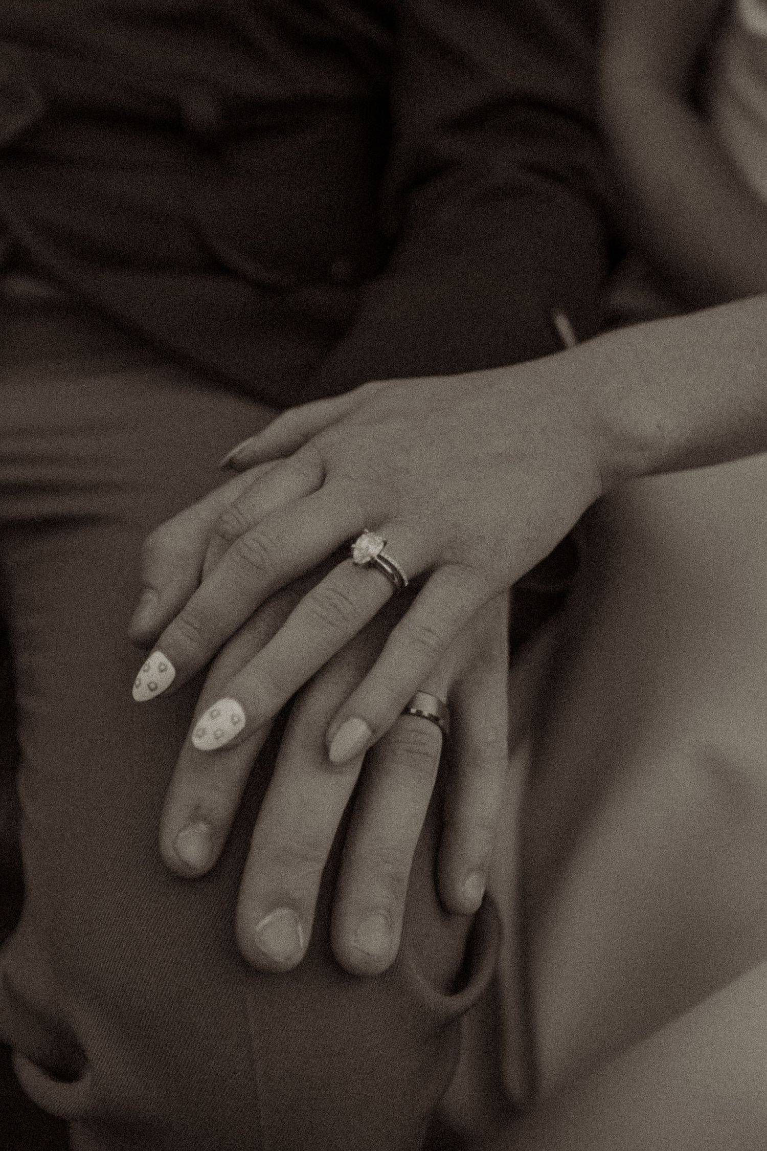 Sepia-toned close-up of two hands resting together, one wearing a diamond engagement ring and decorated nails.