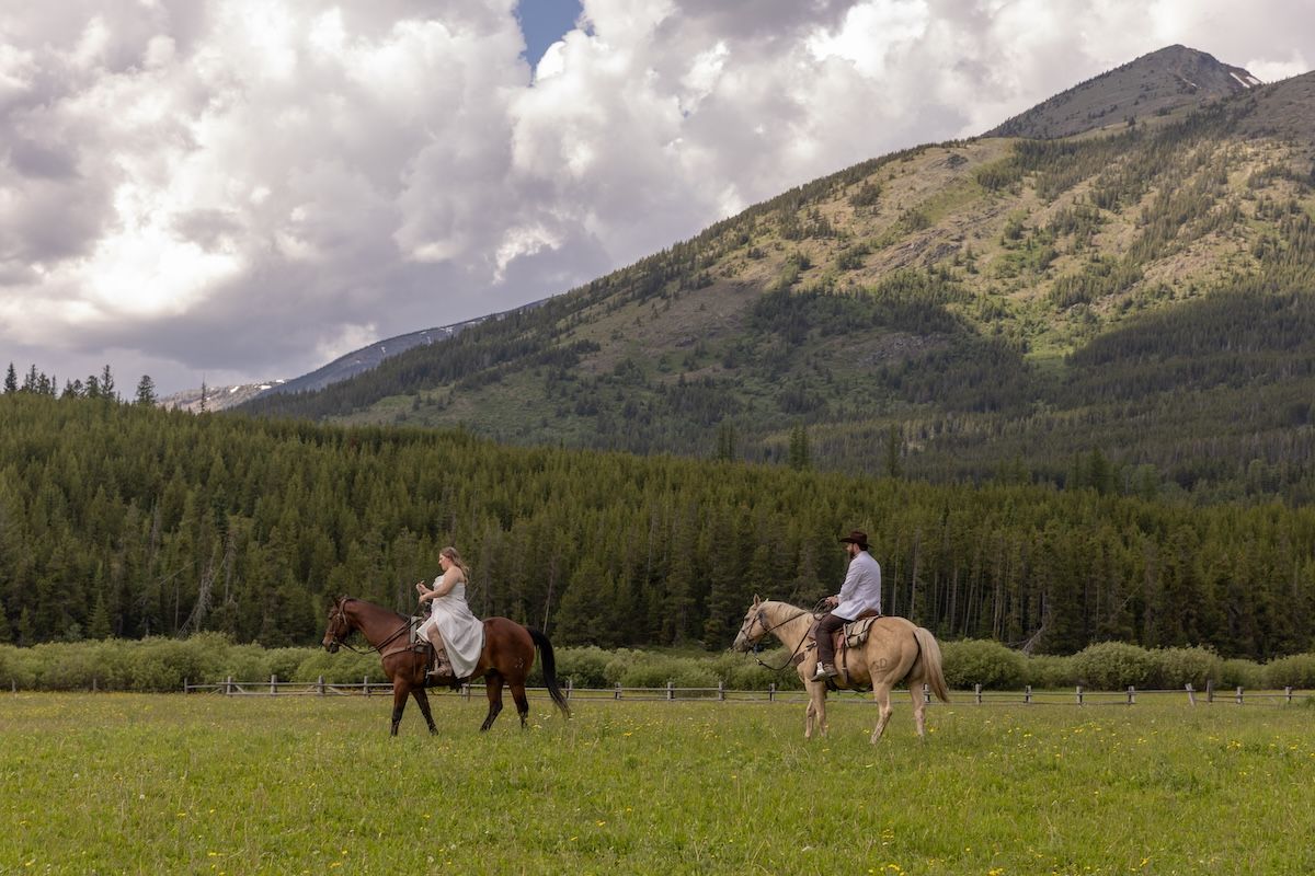 Two riders on horseback cross a lush green meadow with dense pine forest and mountain peaks under cloudy skies.