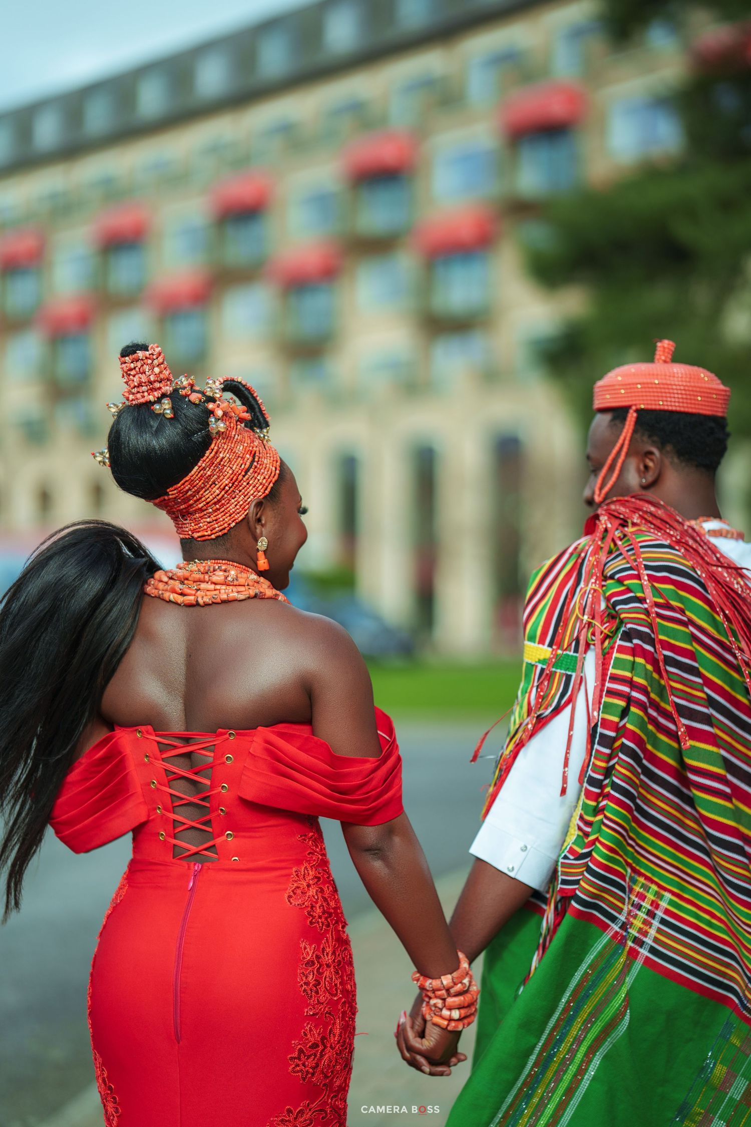 African couple in traditional red and green attire holding hands, walking toward a building.