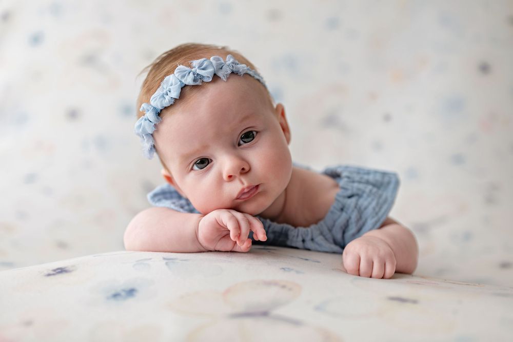Adorable newborn baby girl in blue romper and floral headband lying on soft pastel backdrop.
