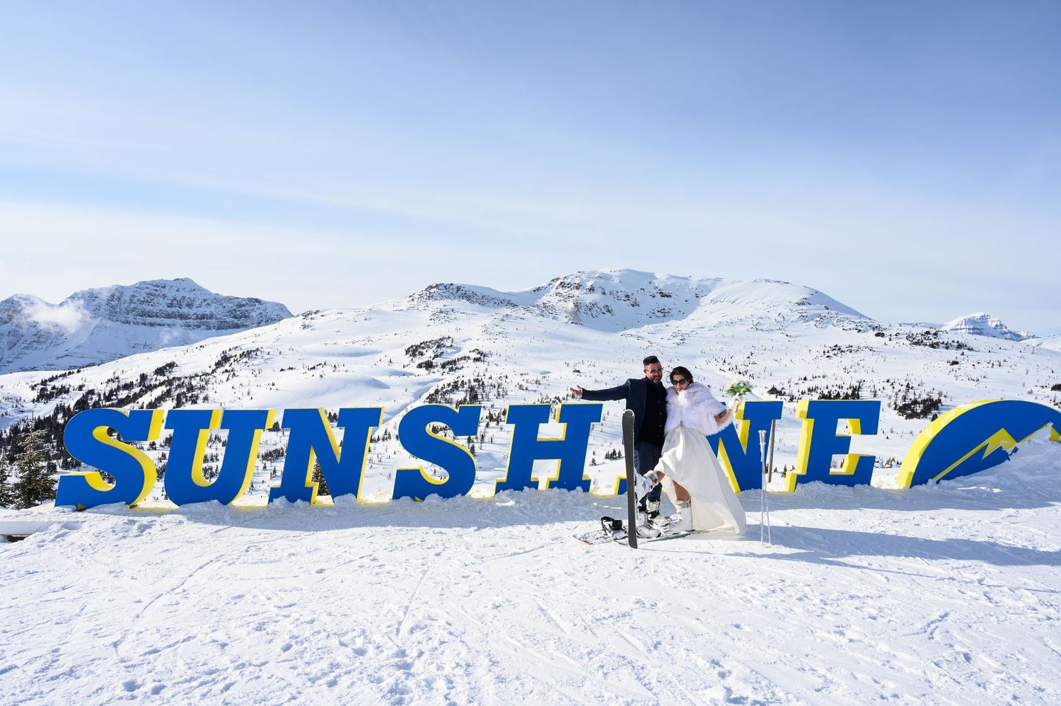 Panoramic view of snowy mountain peaks and ski slopes with a colorful resort sign visible against a bright blue sky.