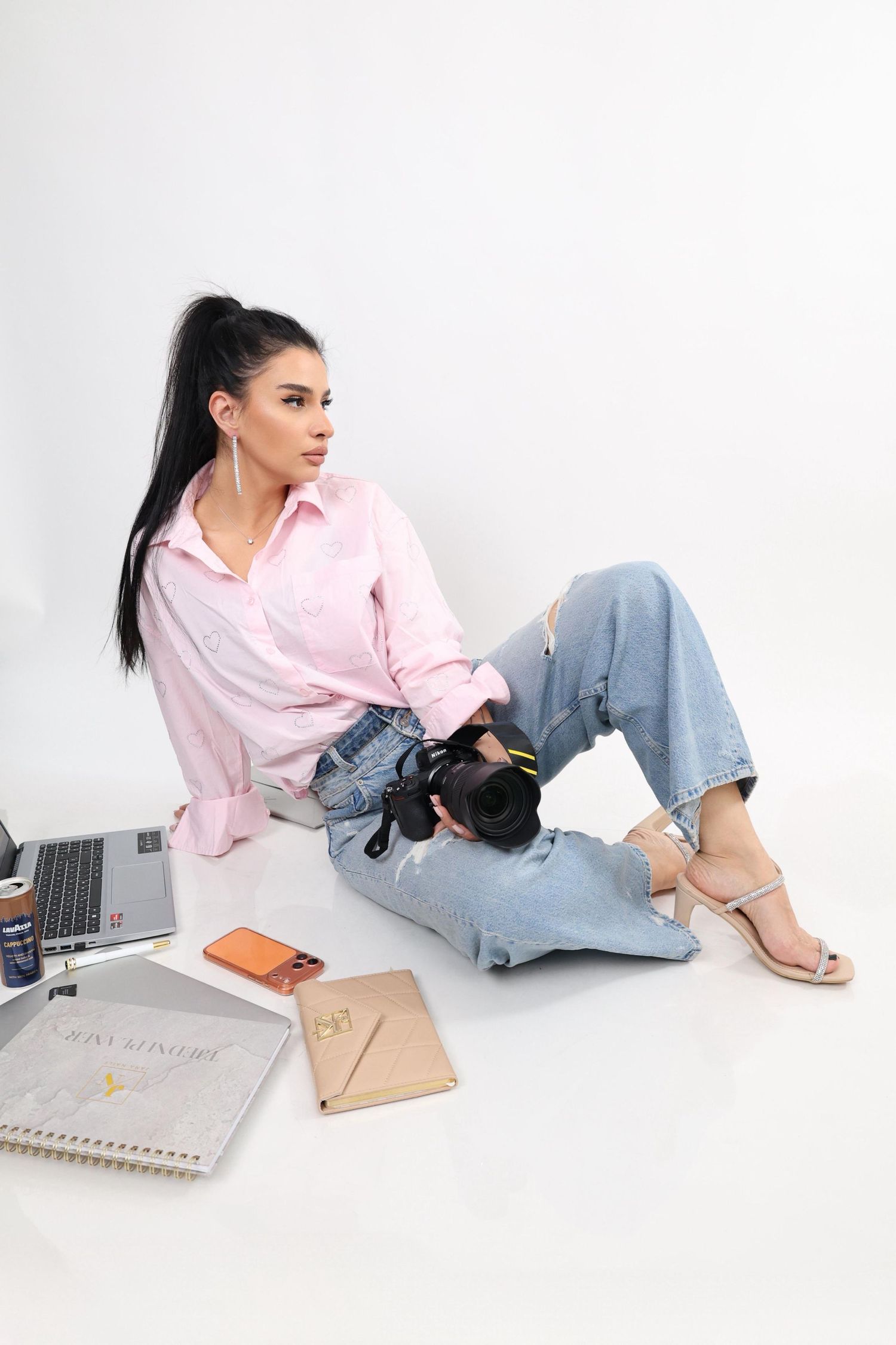 Young woman in pink shirt and jeans sitting on white floor holding camera with laptop and notebooks nearby.