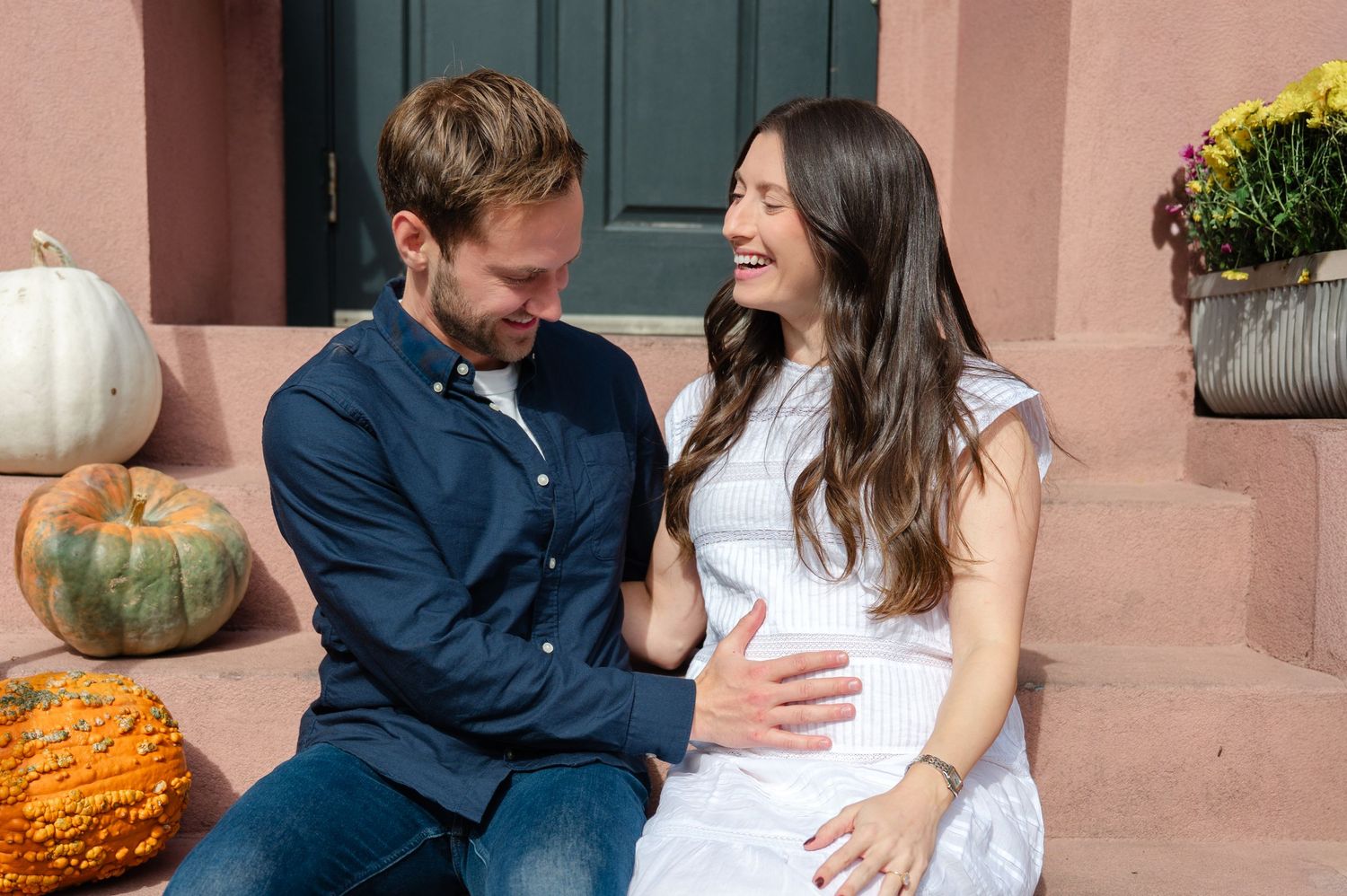 Happy couple sitting on steps with pumpkins, woman is pregnant, smiling at each other in a fall outdoor setting.