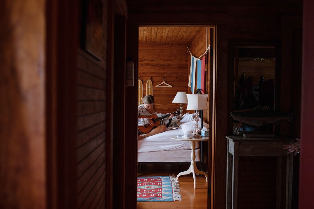 Cozy wooden cabin bedroom viewed through doorway of teen girl playing guitar. Lakes Region NH Southern Maine family vacation photographer.