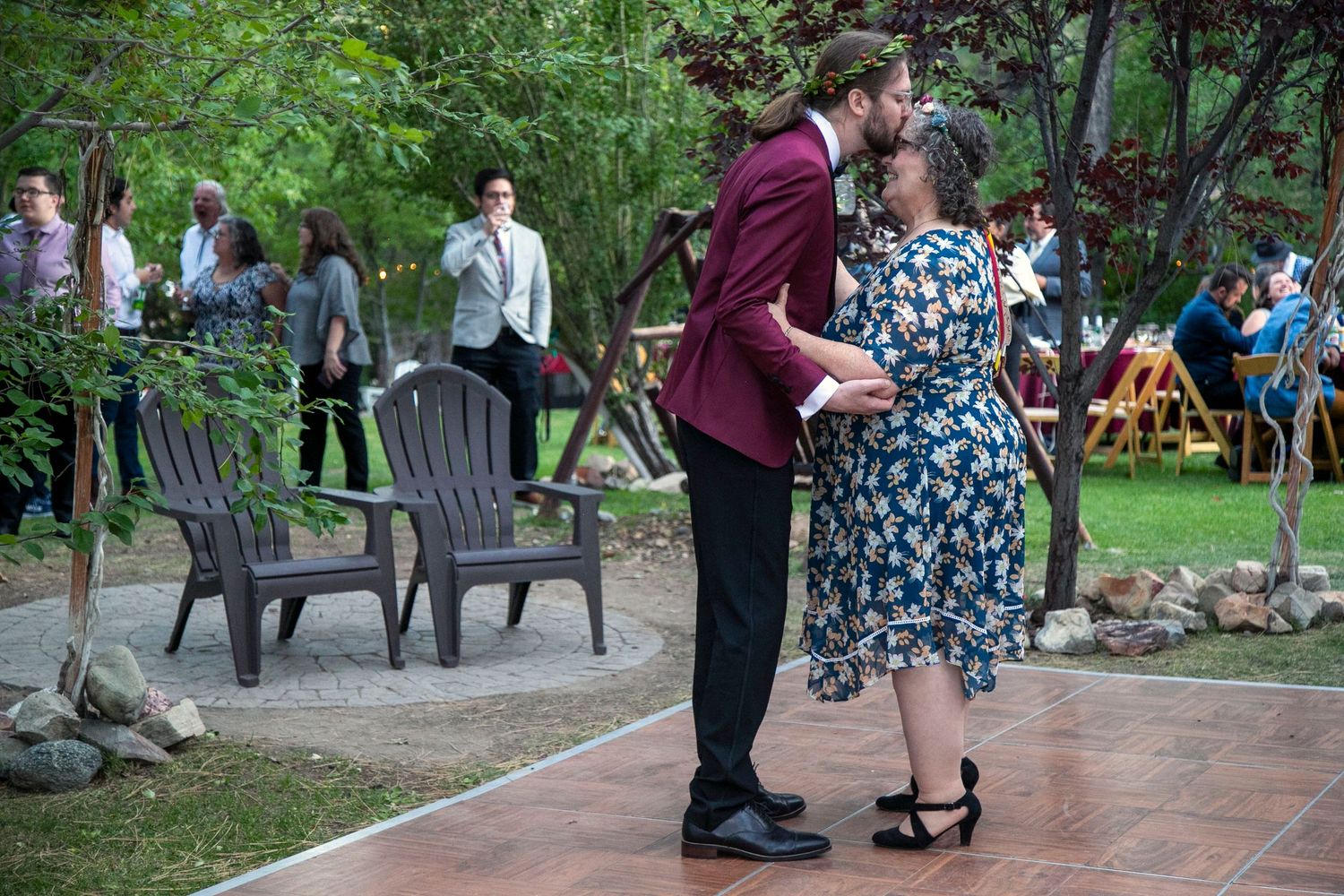 Guests gather outdoors watching two men greet near colorful rugs at an informal celebration in a wooded setting.