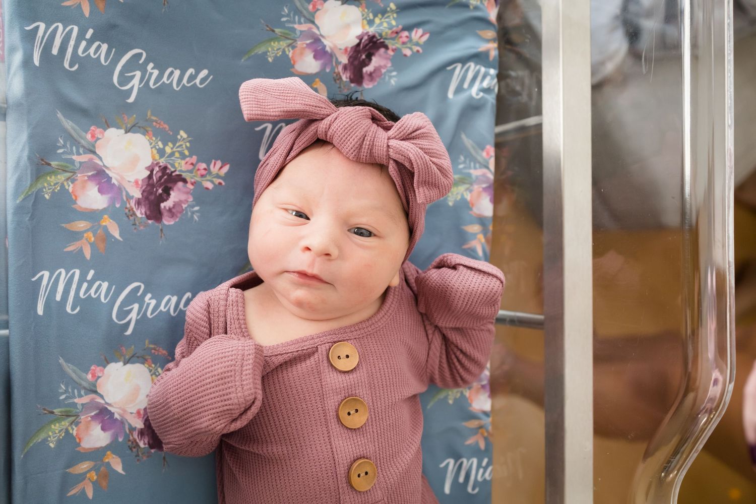 Newborn baby girl named Mia Grace in pink outfit and bow headband lying on personalized floral name blanket.