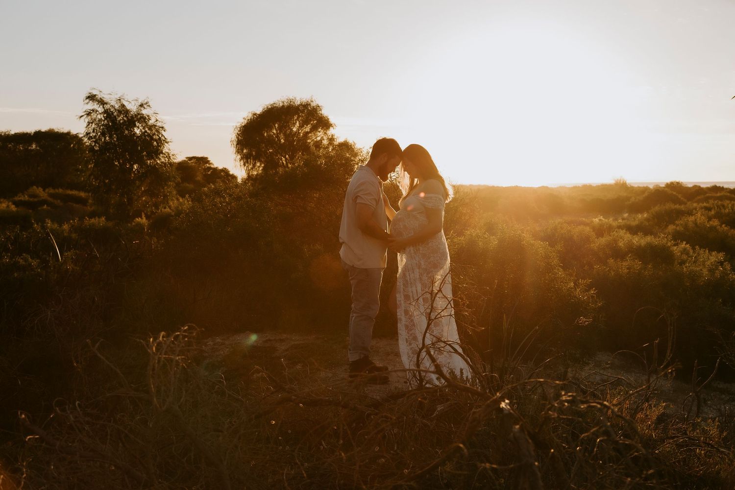 Couple silhouetted against golden sunset light in a natural landscape, sharing an intimate moment surrounded by lush greenery.