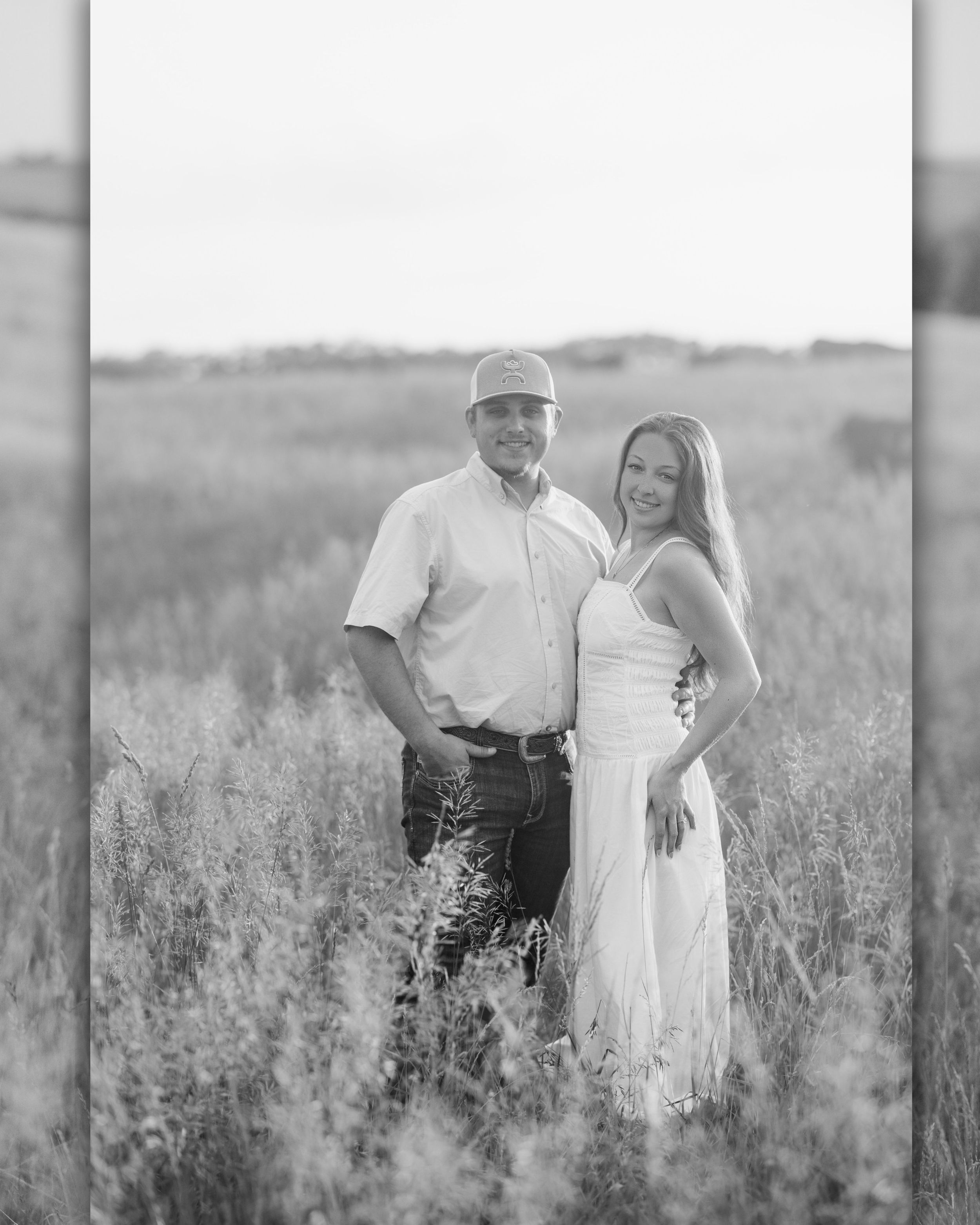 Black and white photo of a couple standing in a field, man in cap and shirt, woman in white dress, smiling outdoors.