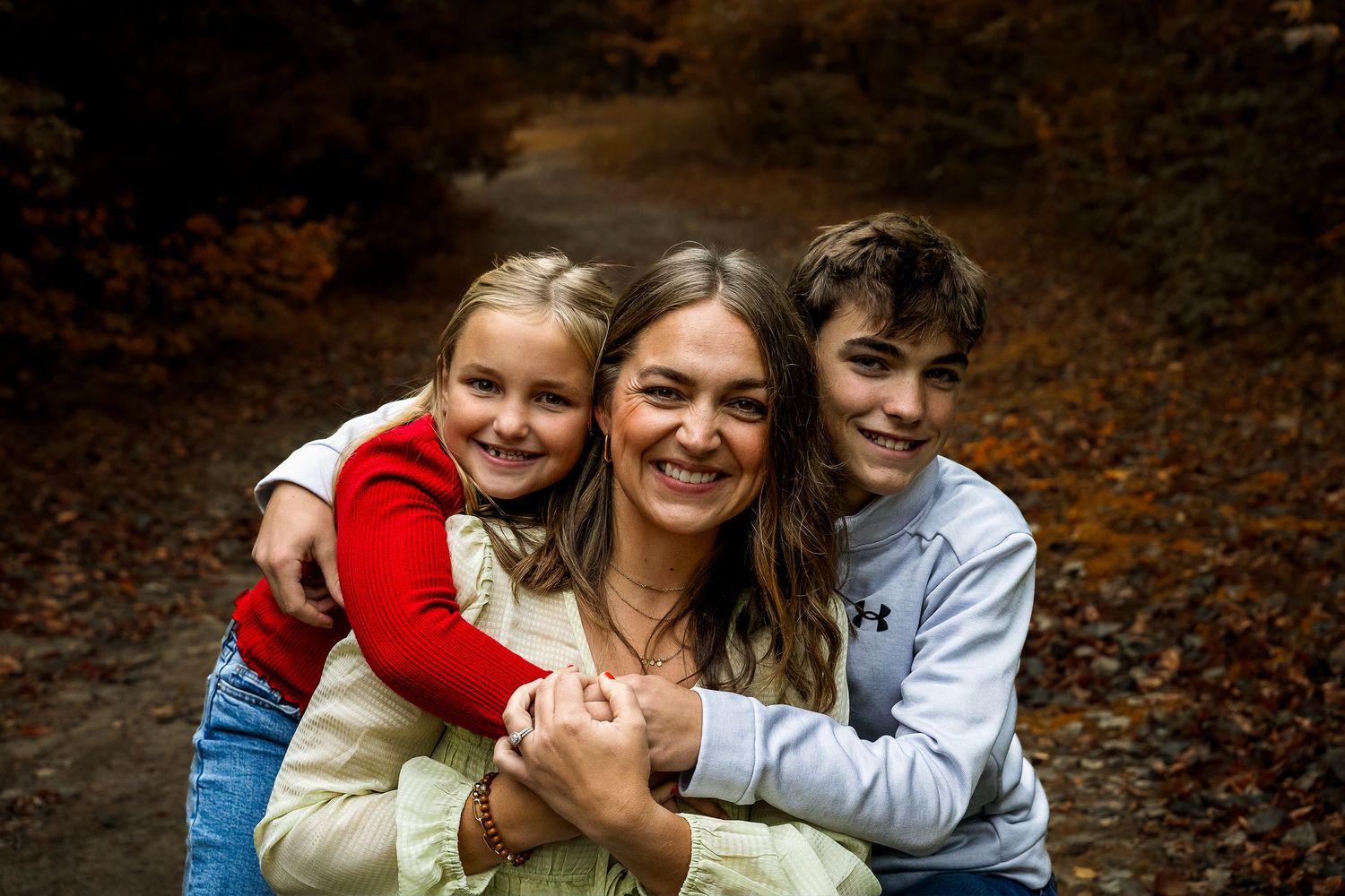 A smiling woman poses with two children hugging her in an autumn forest setting with colorful fall foliage.
