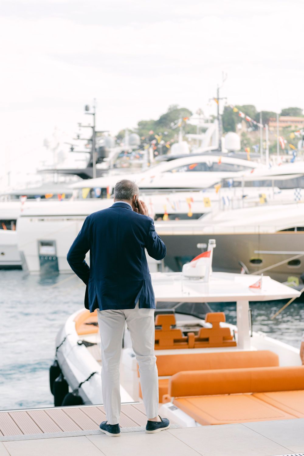 Man in navy blazer and white pants stands on marina dock talking on phone with luxury yachts in background.