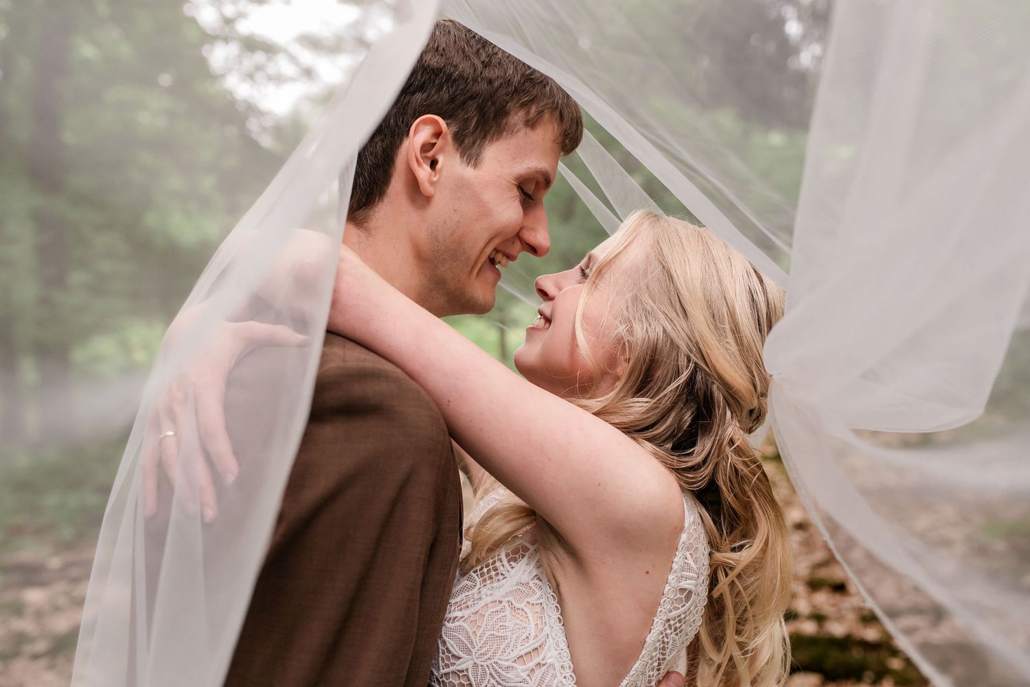 Bride and groom share an intimate moment under a flowing veil in a misty forest setting.