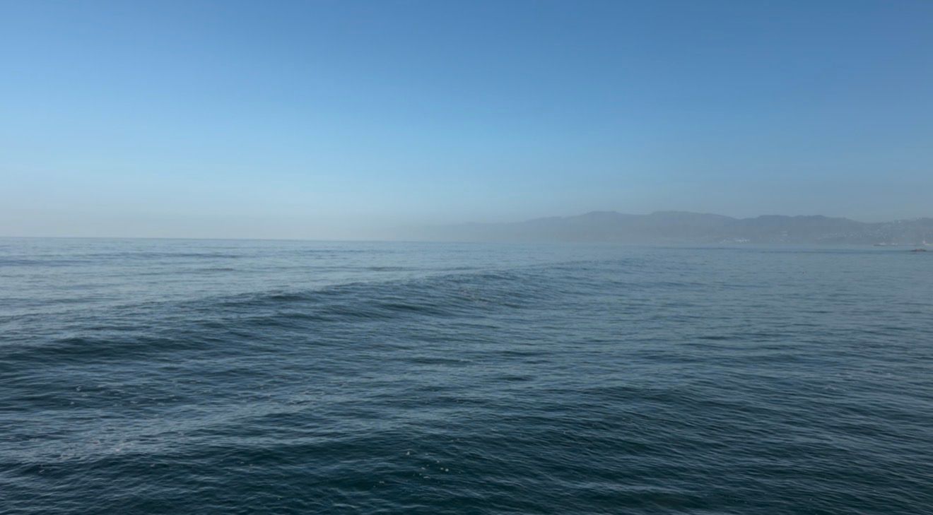 Calm ocean waves with a hazy mountain coastline on the horizon under a clear blue sky.