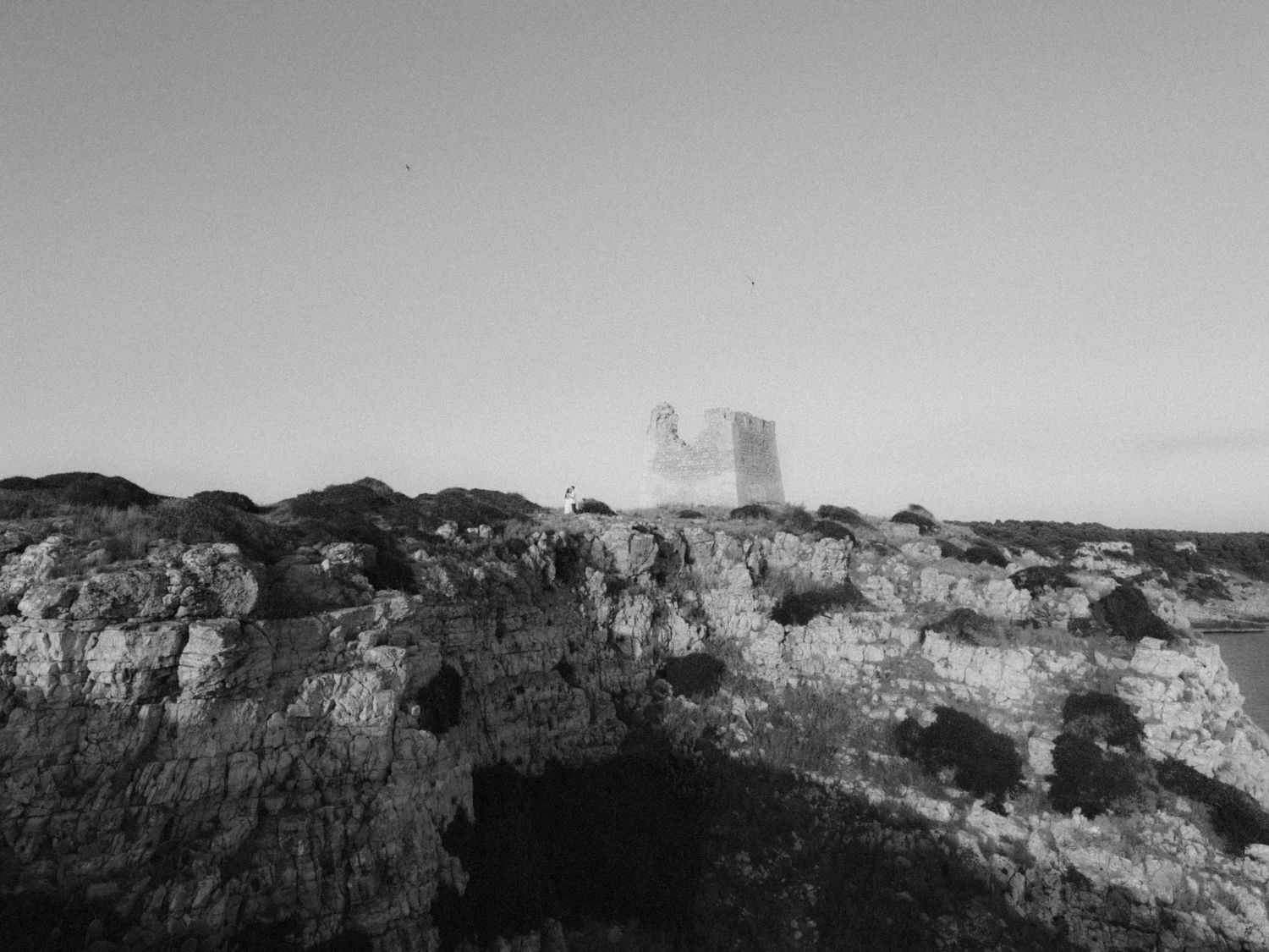 Black and white photo of ancient rocky coastline with a historic stone tower ruins on the horizon.