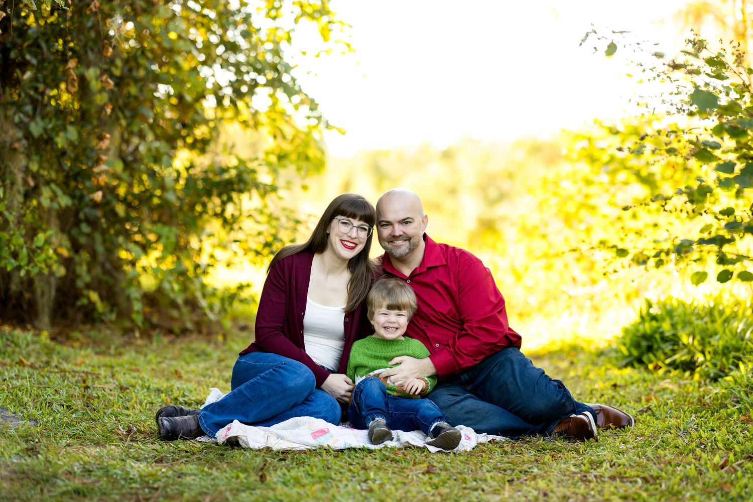 Family of three smiling and sitting on grass in a sunny outdoor park setting with lush green trees in background.