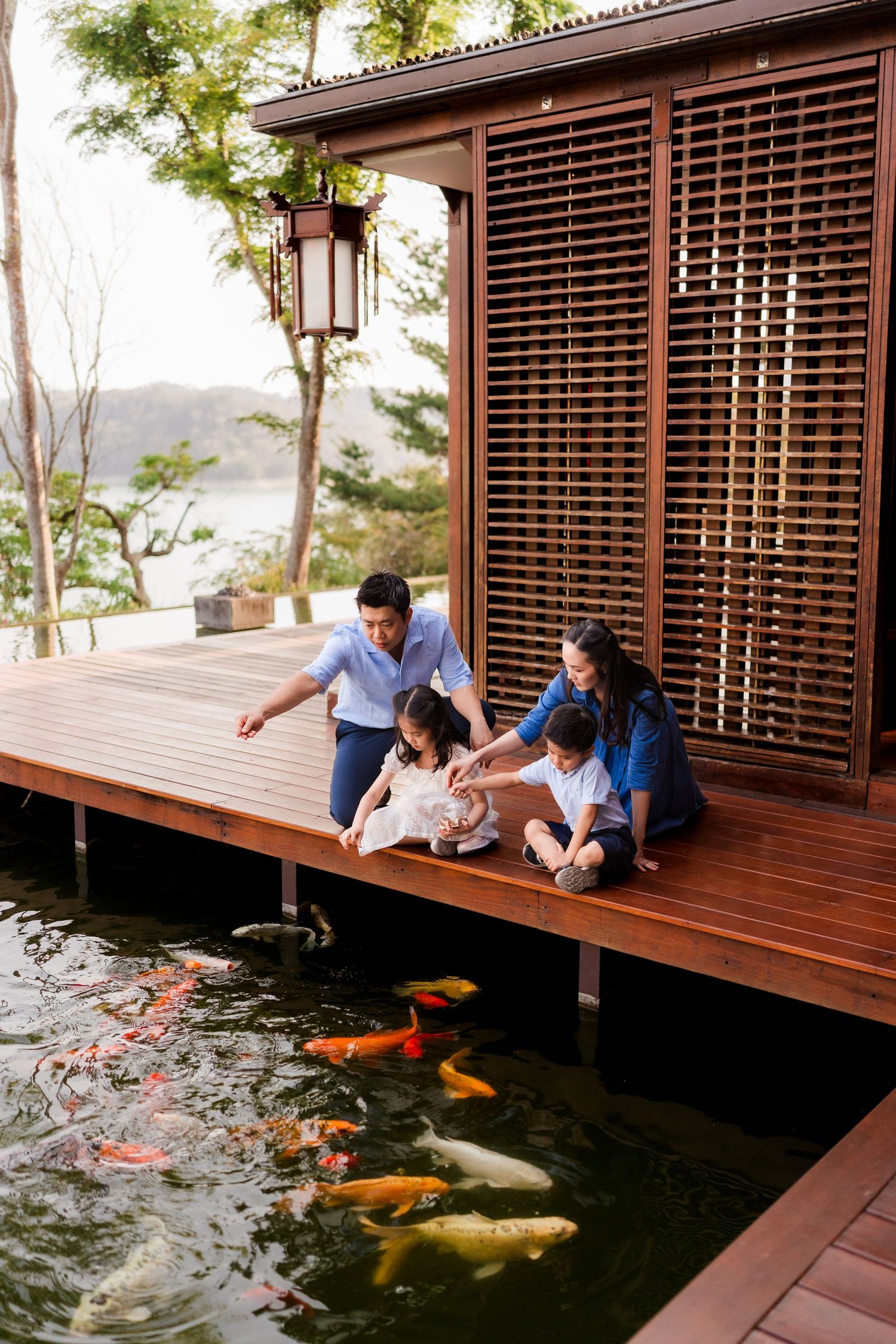 Family feeding colorful koi fish from a wooden deck beside a traditional Japanese-style building.