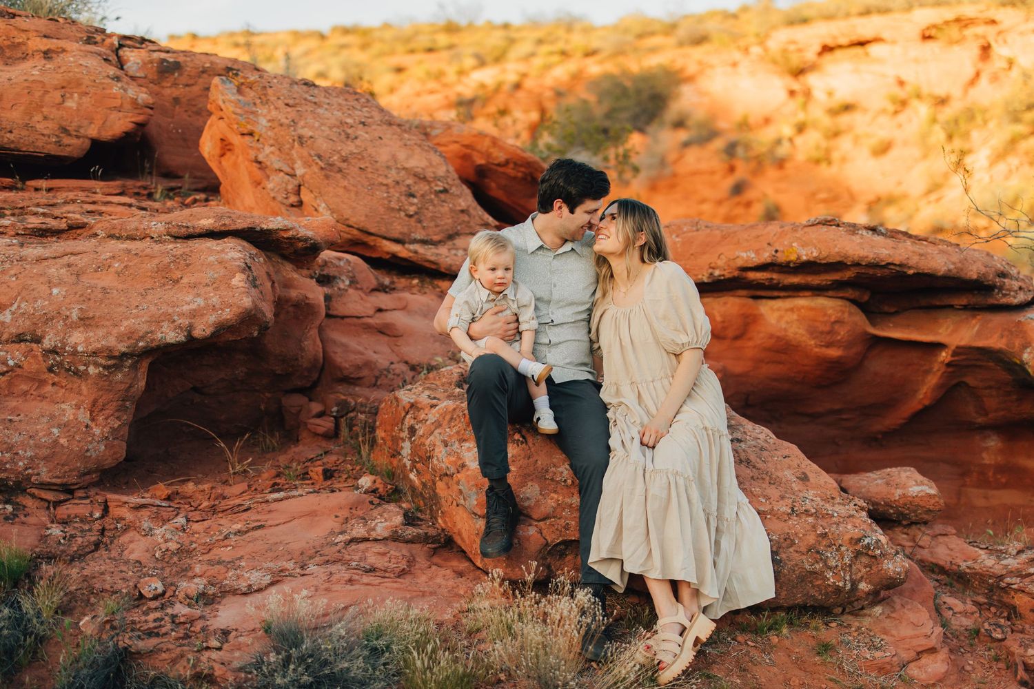 A couple standing on a rocky desert landscape, with the woman wearing a long dress and the man holding a young child.