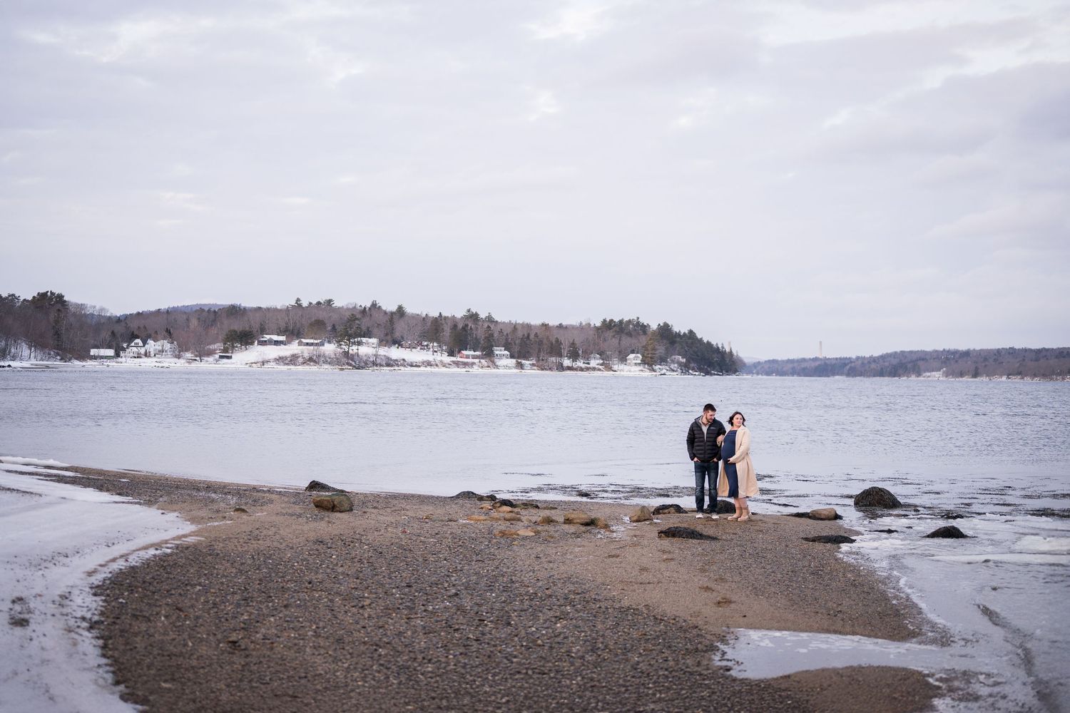 A couple shares a tender moment on a snowy winter beach with wooden pilings and water in the background.