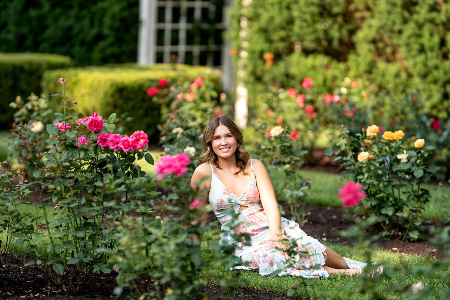 Woman in floral dress sitting among colorful roses in a lush garden, surrounded by pink and yellow blooms.